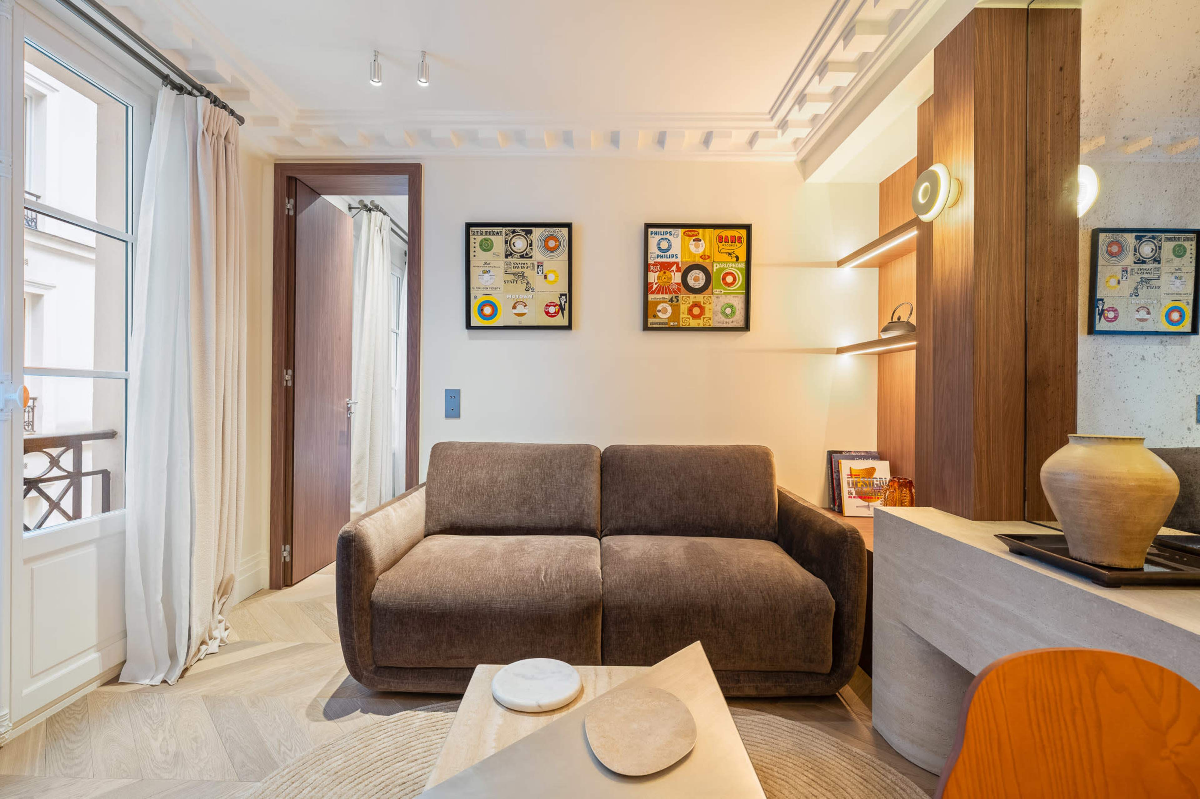 The image shows a modern living space featuring a brown sofa, a wooden accent wall, framed art, and a small table with decorative items.