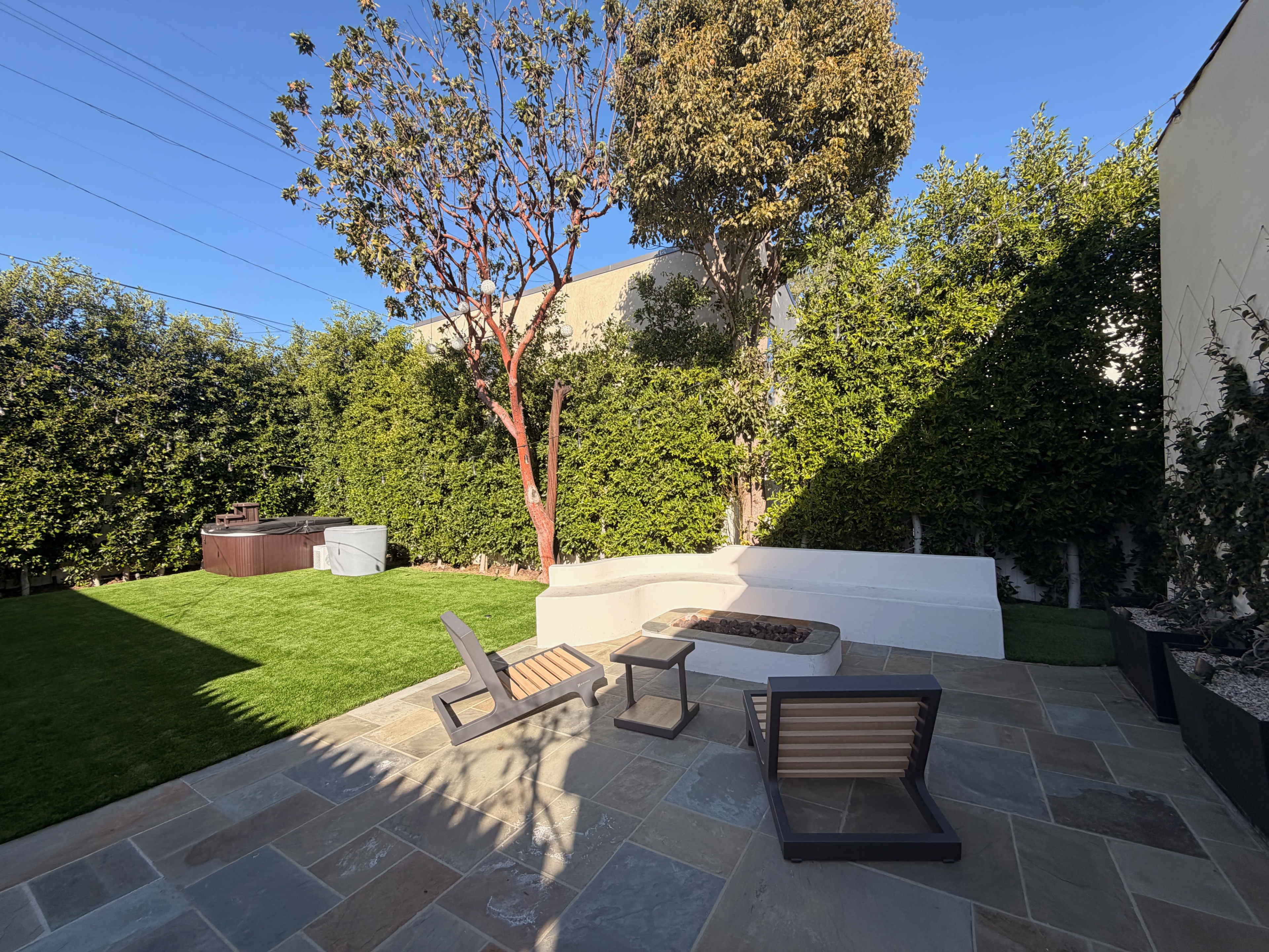 The image shows a landscaped backyard featuring a seating area with two chairs, a fire pit, a grassy lawn, and a hot tub surrounded by tall greenery.