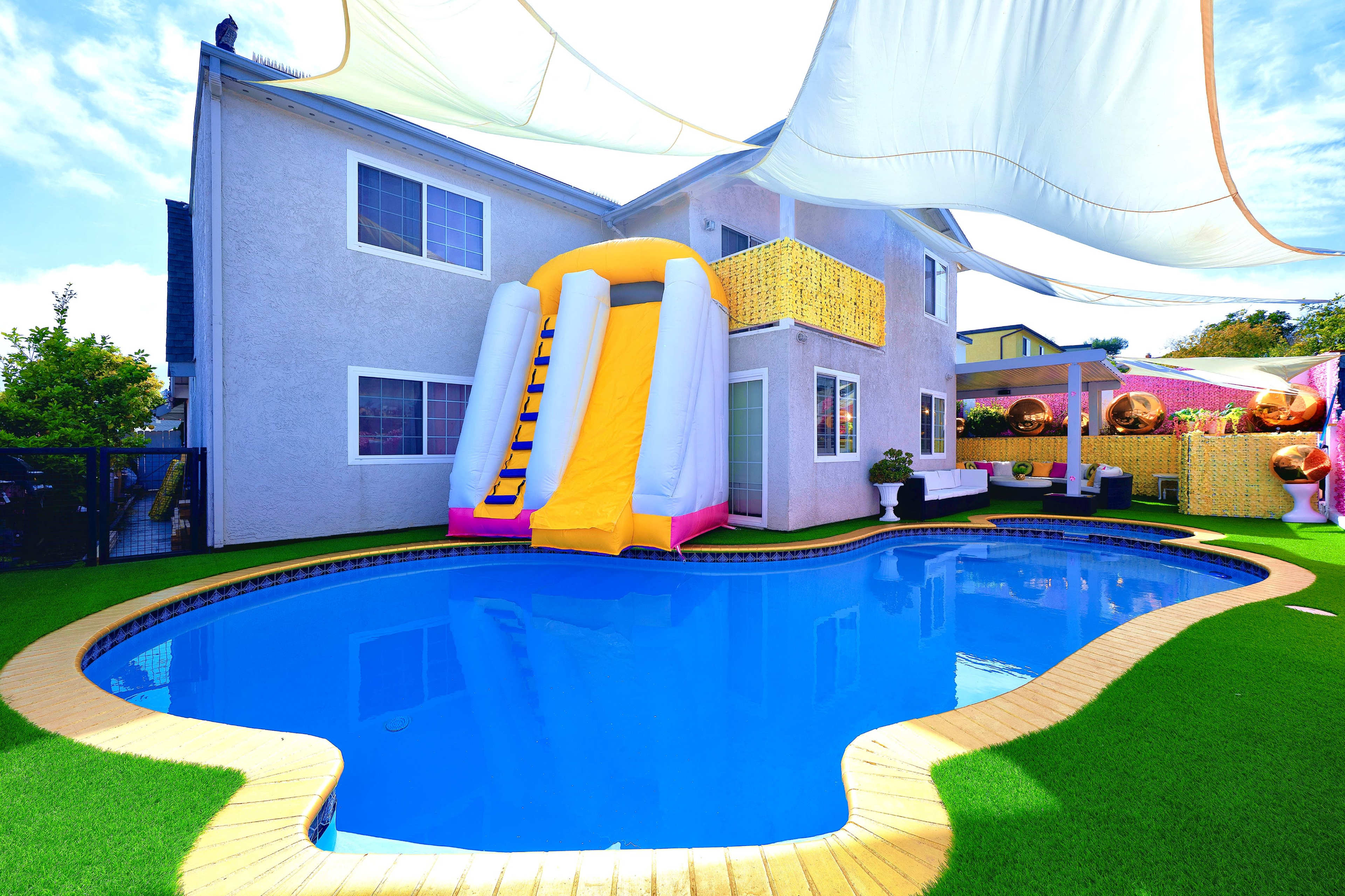 A bright and colorful backyard features a blue swimming pool with a yellow and pink water slide and shaded areas.