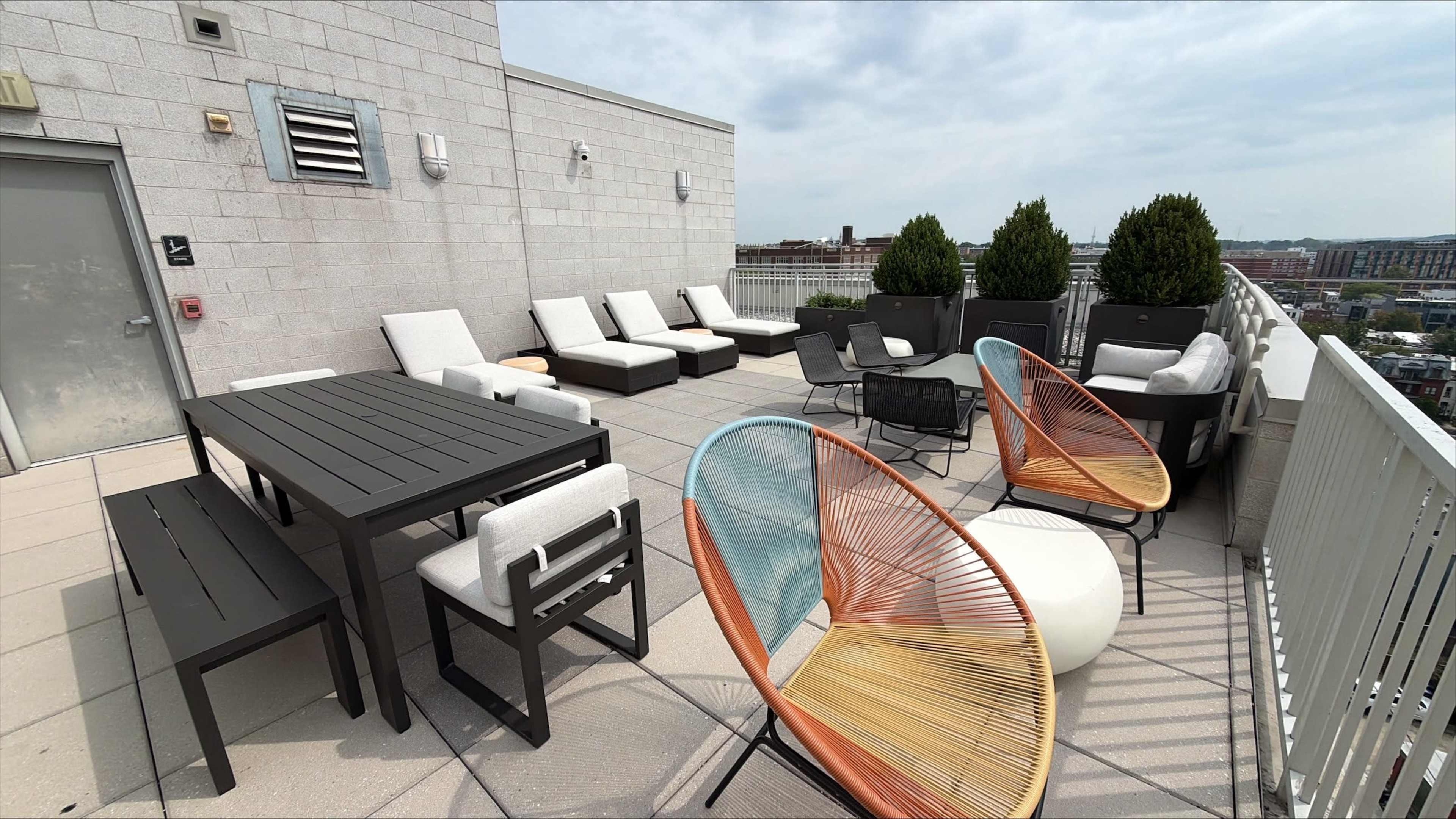 The rooftop terrace features modern lounge furniture, including a dining table, sun loungers, and colorful chairs, with potted plants lining the edges.