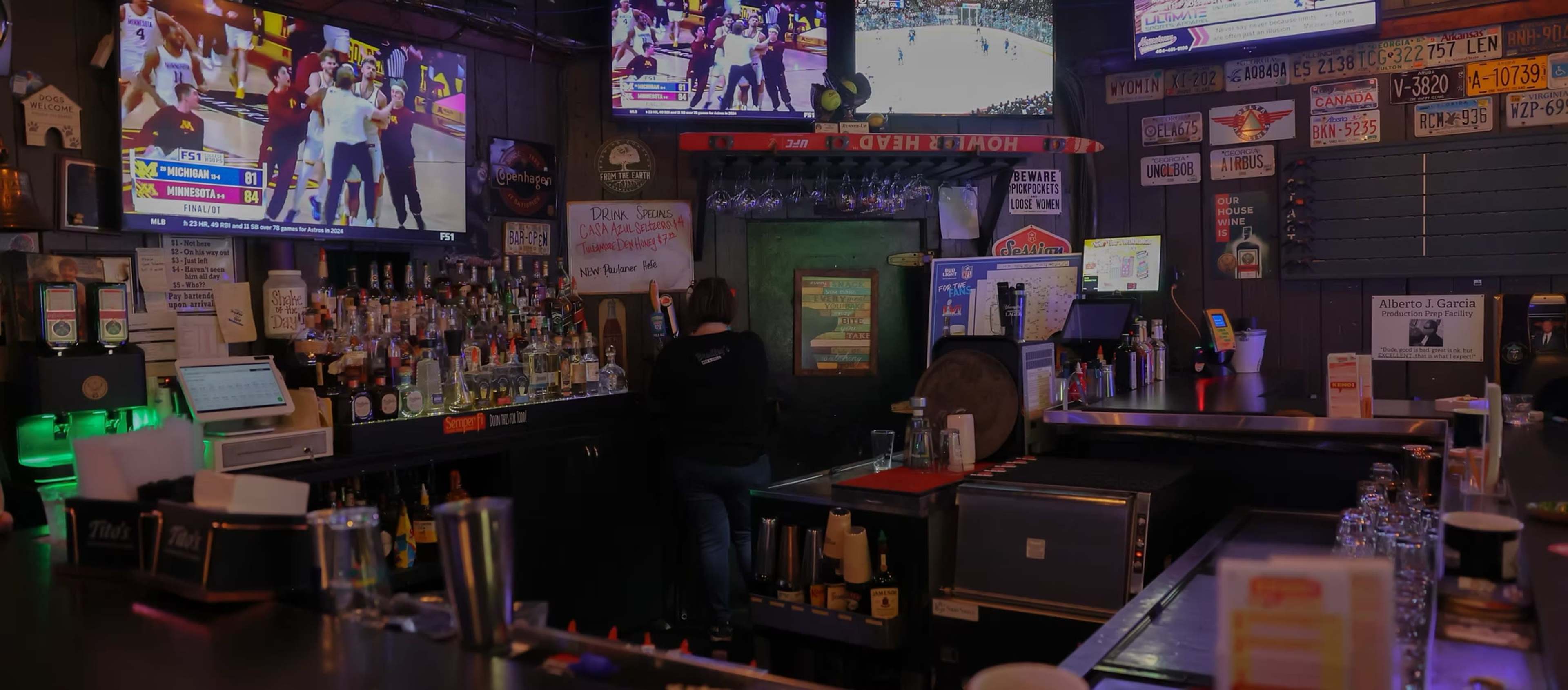 The image depicts a lively bar scene with multiple screens showing sports events and a bartender preparing drinks at the counter.