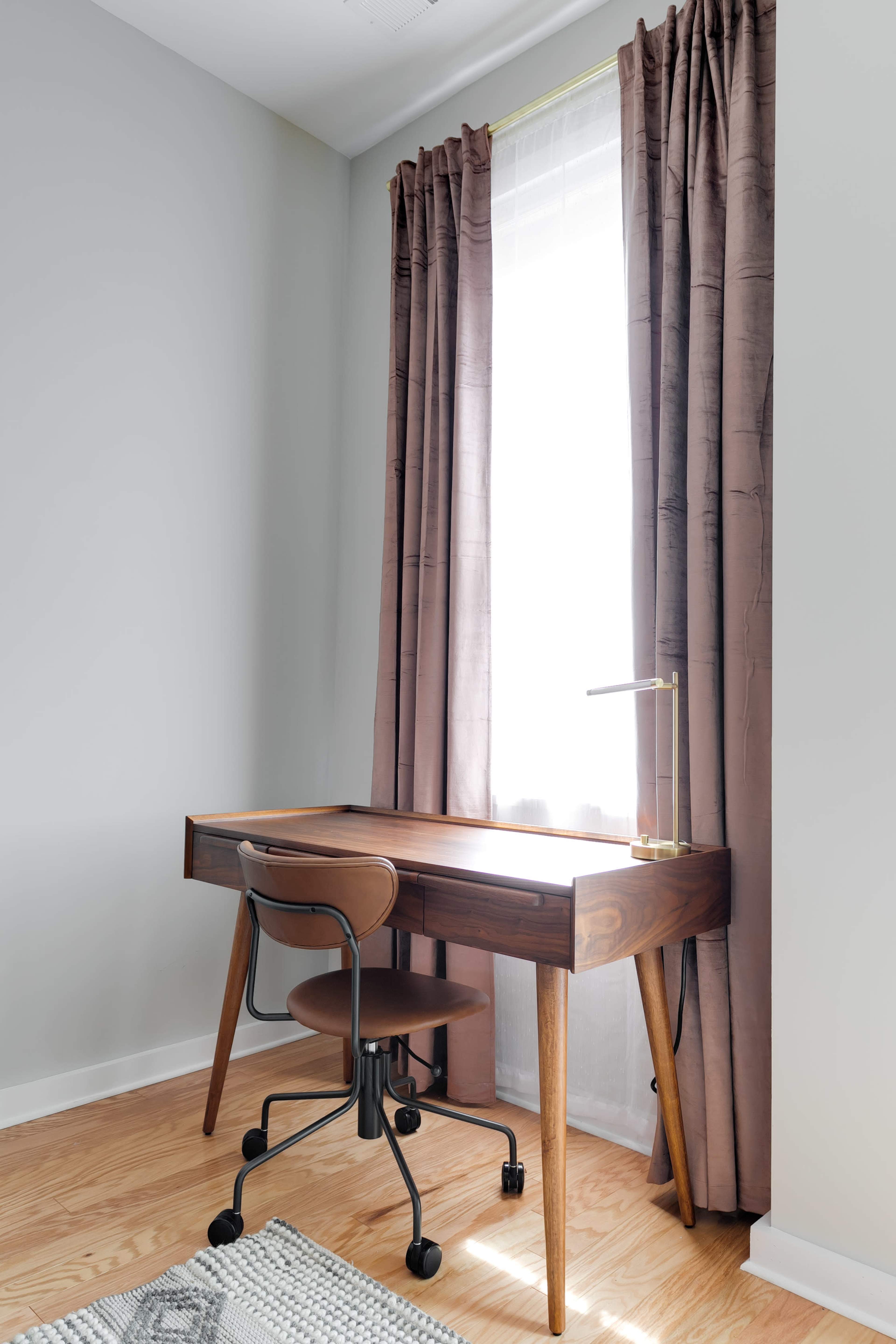 A modern wooden desk with a chair is positioned beside a large window covered with brown curtains in a well-lit room.