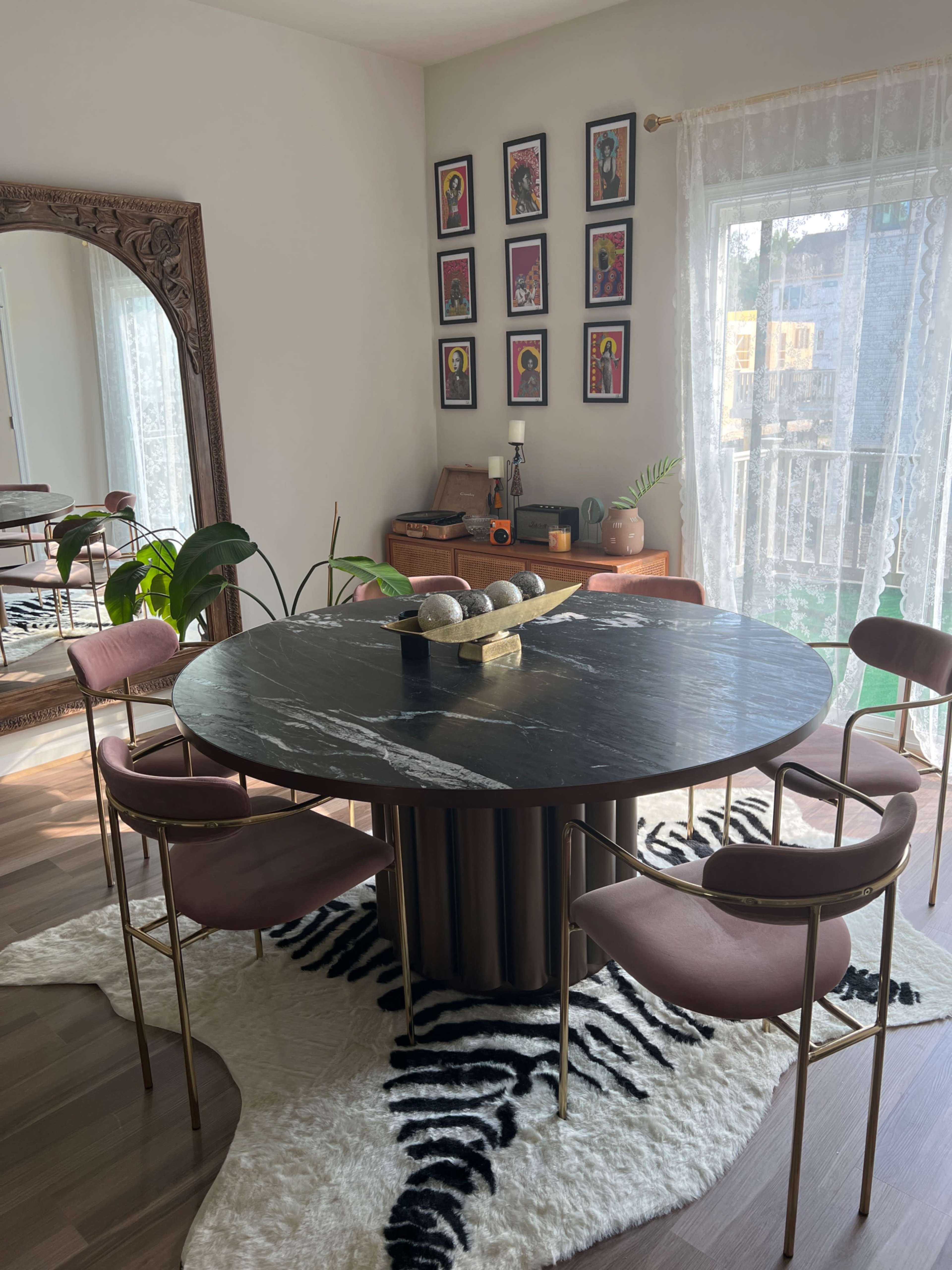 A circular marble dining table surrounded by four pink chairs, situated on a zebra-patterned rug in a well-lit room decorated with framed artwork on the walls.