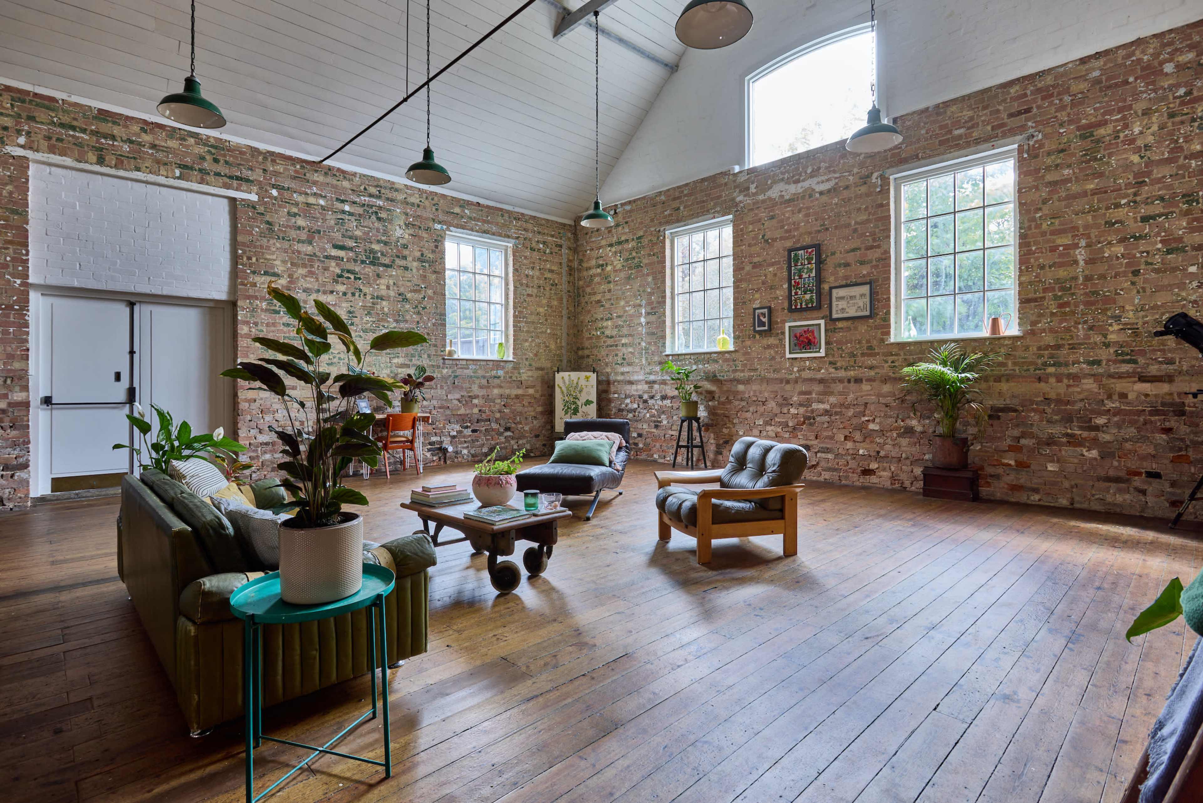A spacious room with exposed brick walls, wooden floors, and large windows, furnished with green sofas, a wooden coffee table, and various plants.