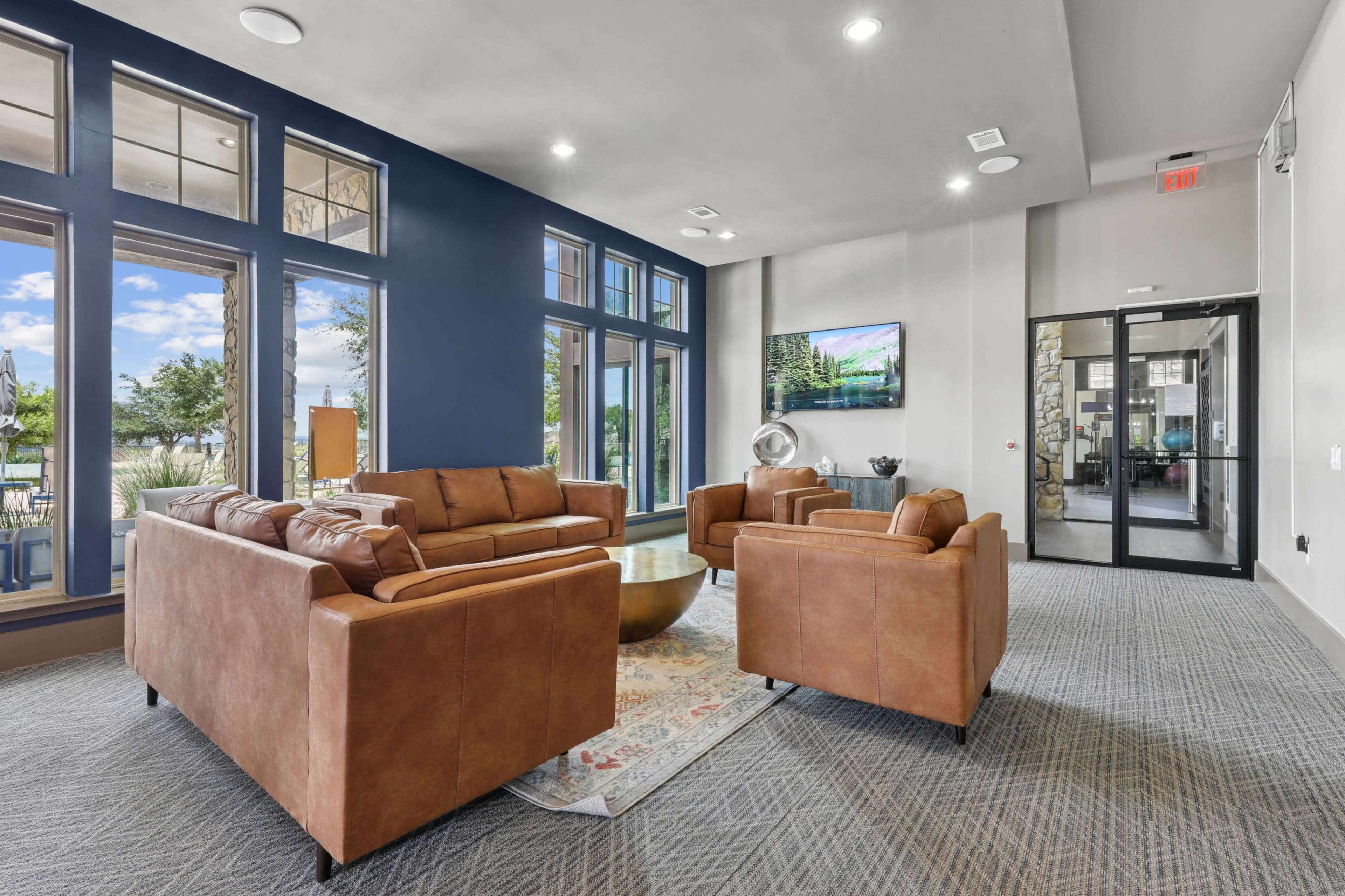 The image shows a modern lounge area with two brown leather sofas, two armchairs, a round coffee table, large windows, and a doorway leading to an outdoor space.