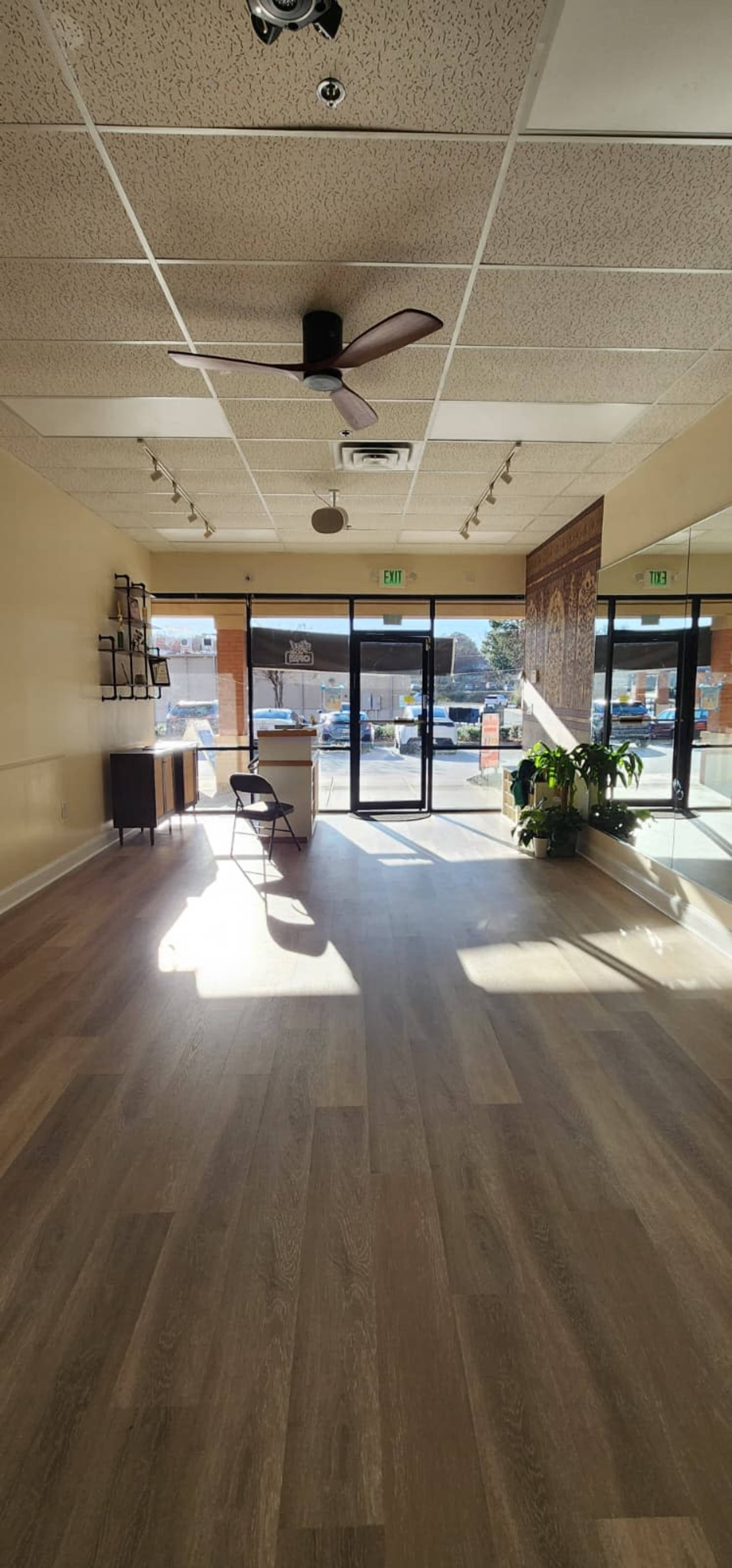The image shows an empty interior of a spacious commercial space with wooden flooring, large windows, and a ceiling fan.