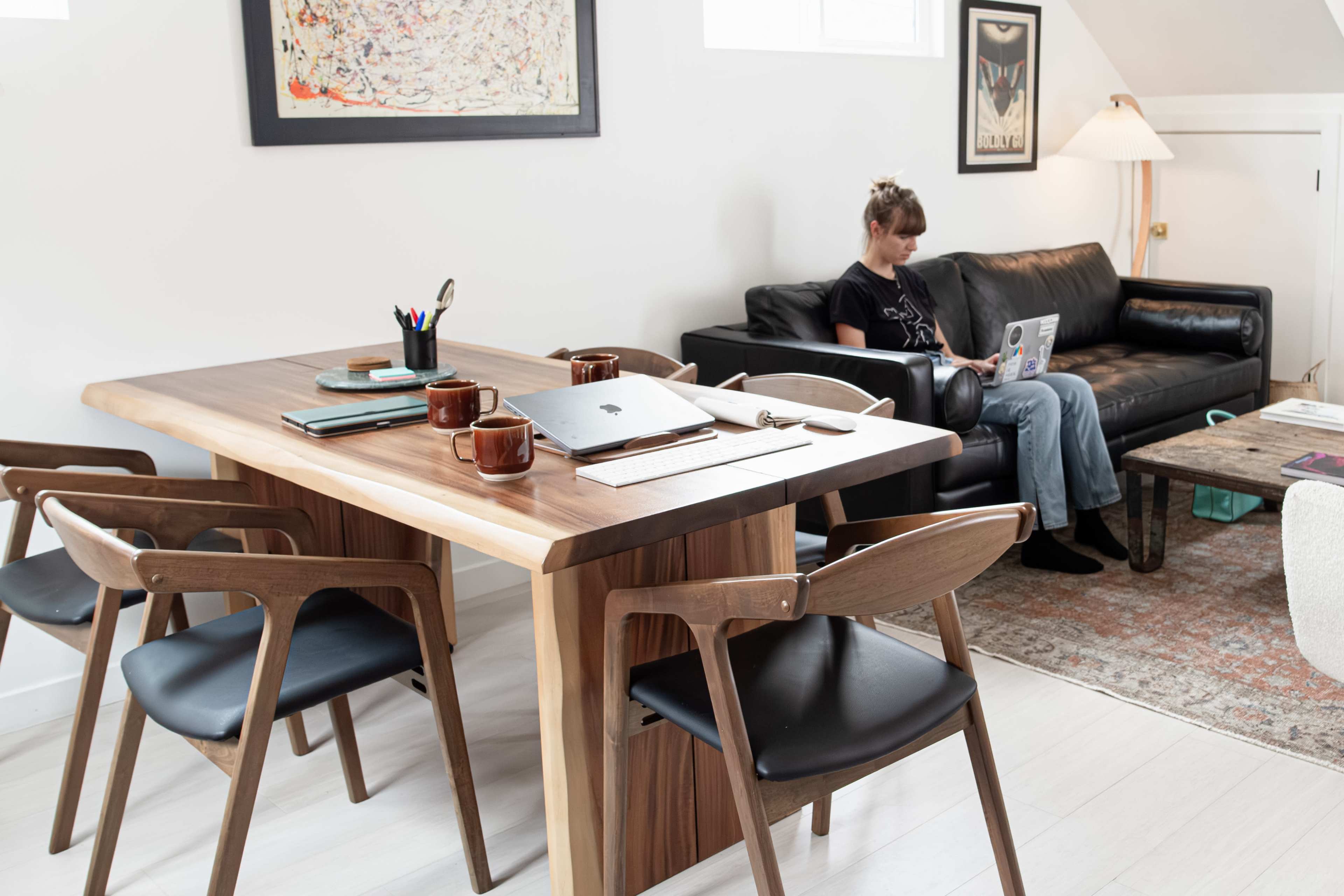 A woman works on a laptop while seated on a couch, and a wooden dining table with chairs and coffee mugs is in the foreground.