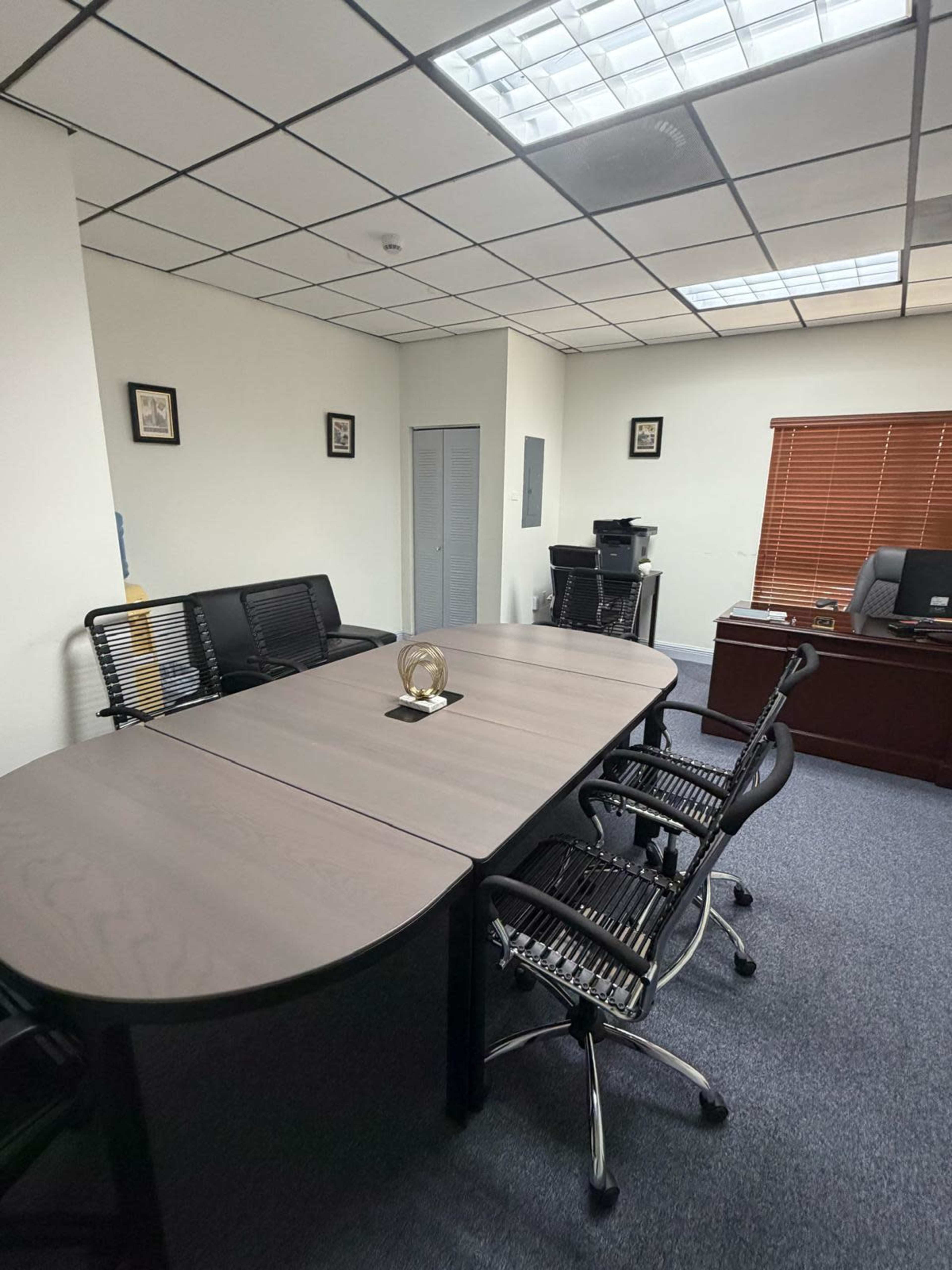 The image depicts a modern office space featuring a large conference table surrounded by black rolling chairs, with a desk and a window covered by blinds in the background.