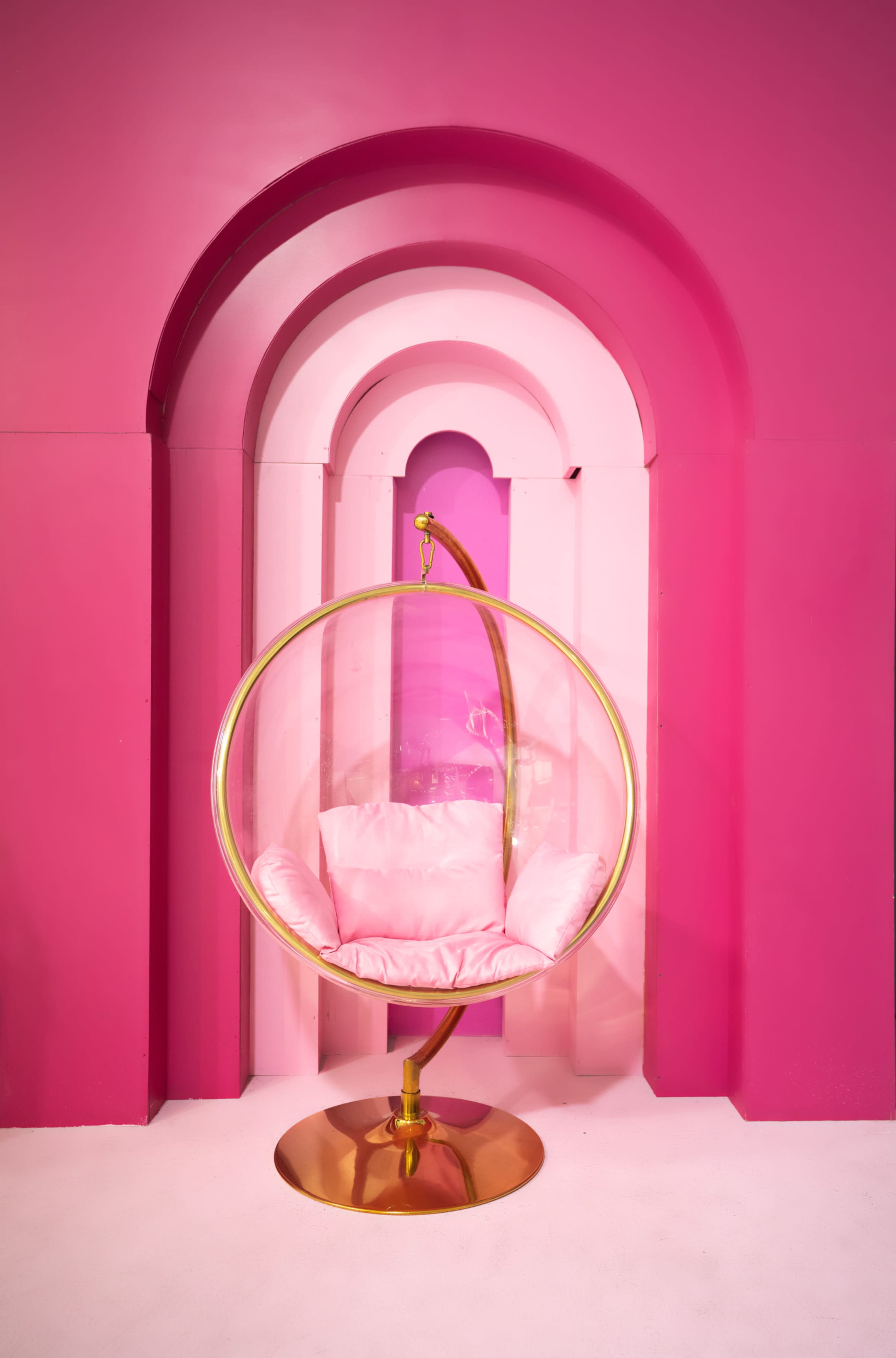 A round, gold chair hangs in a space featuring bright pink walls and arches.