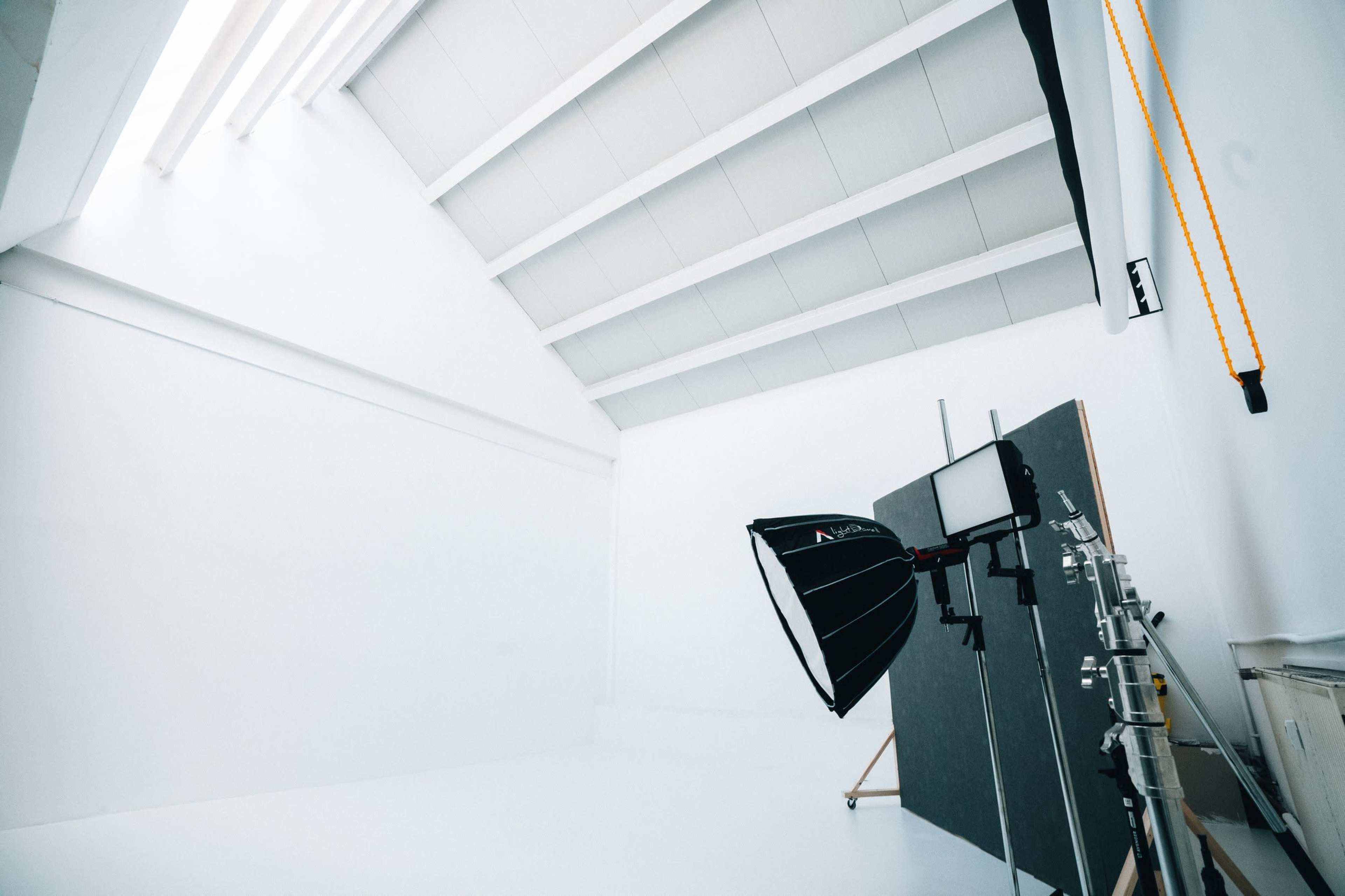 A photography studio features a large white wall, overhead lighting, and equipment including a softbox and a light stand.