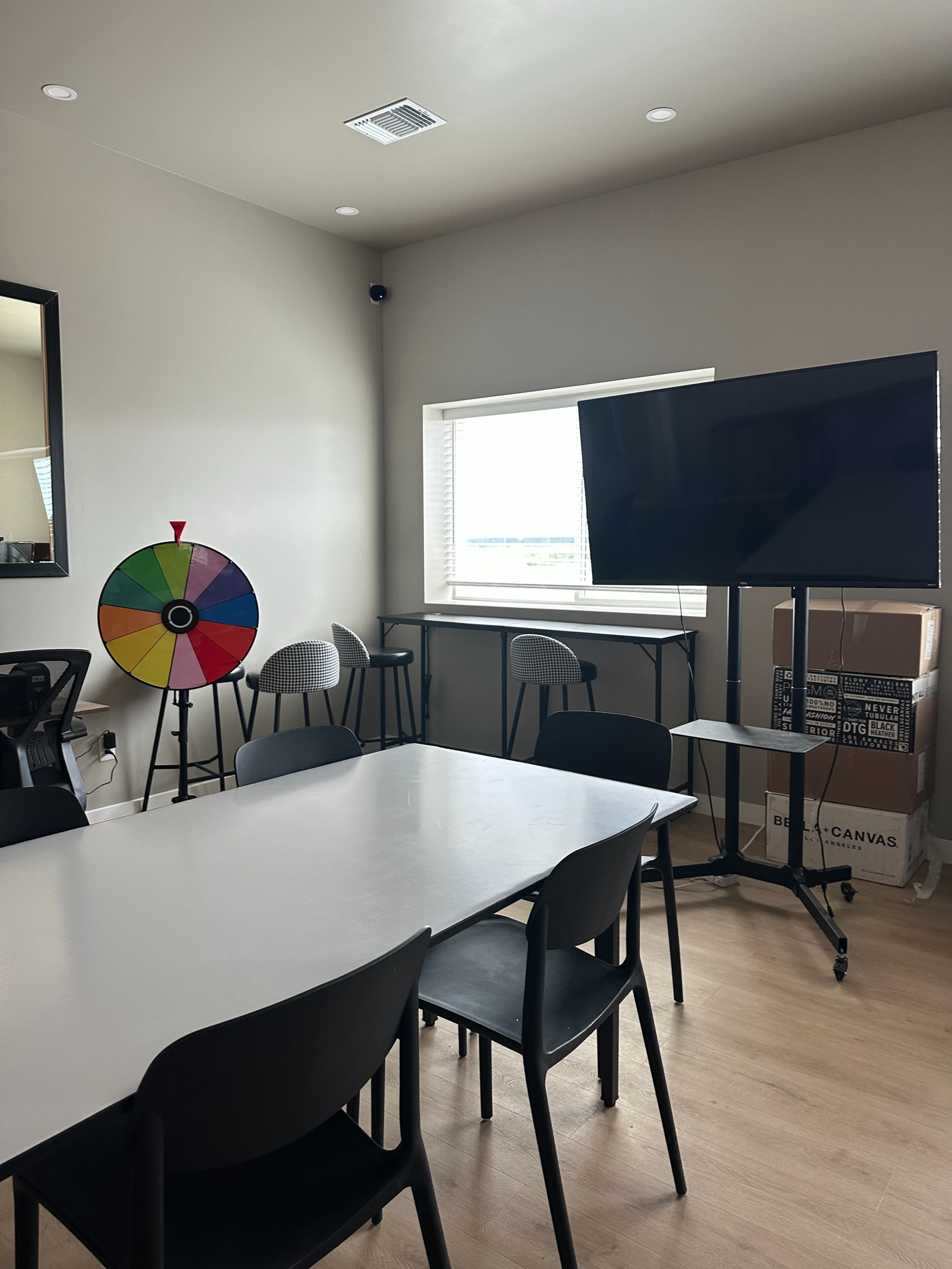 The image shows a meeting room with a large table, several chairs, a colorful spinning wheel, and a flat-screen TV mounted on a stand.