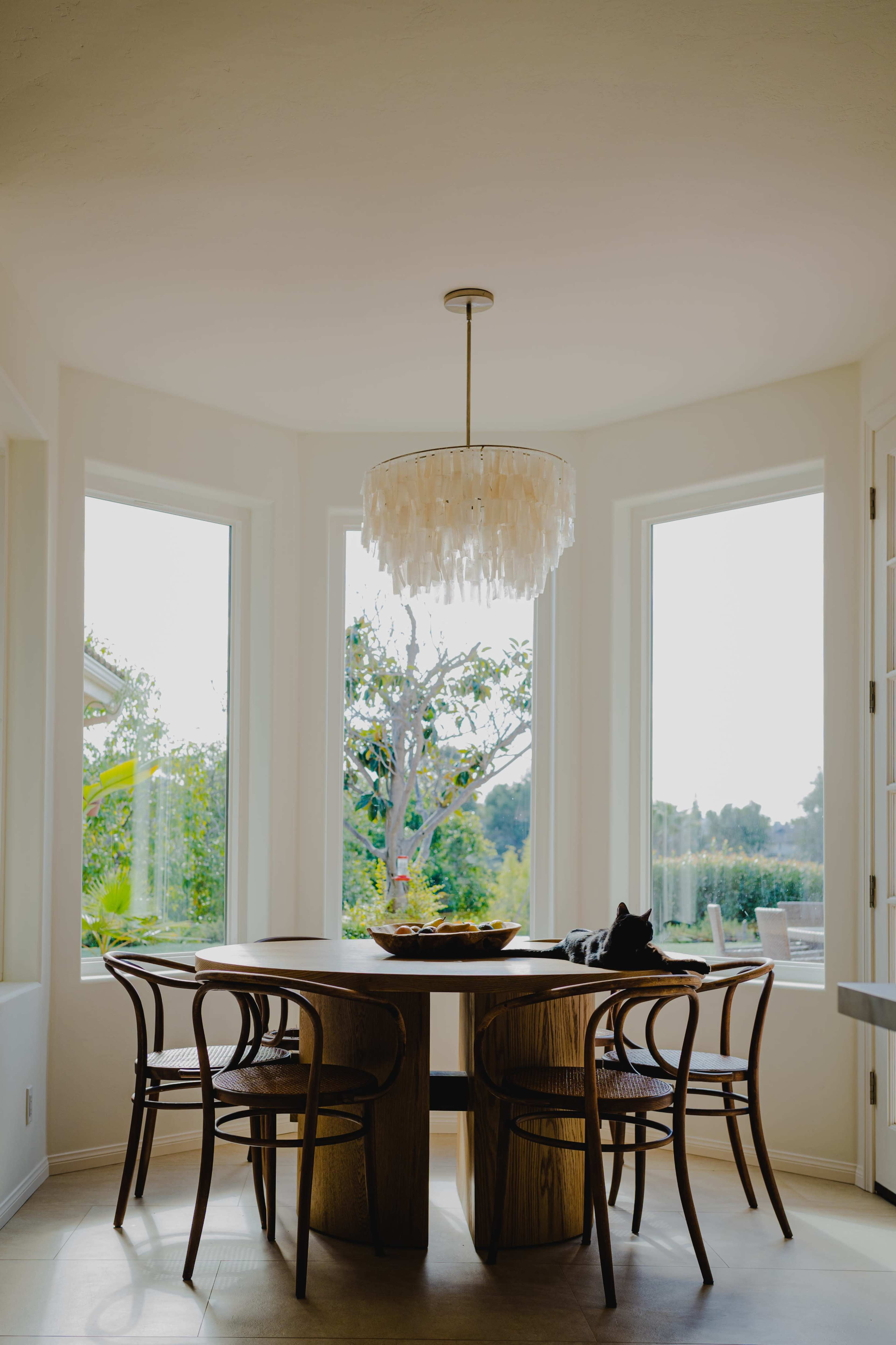 A dining area features a circular wooden table surrounded by four chairs, with large windows providing a view of greenery outside.
