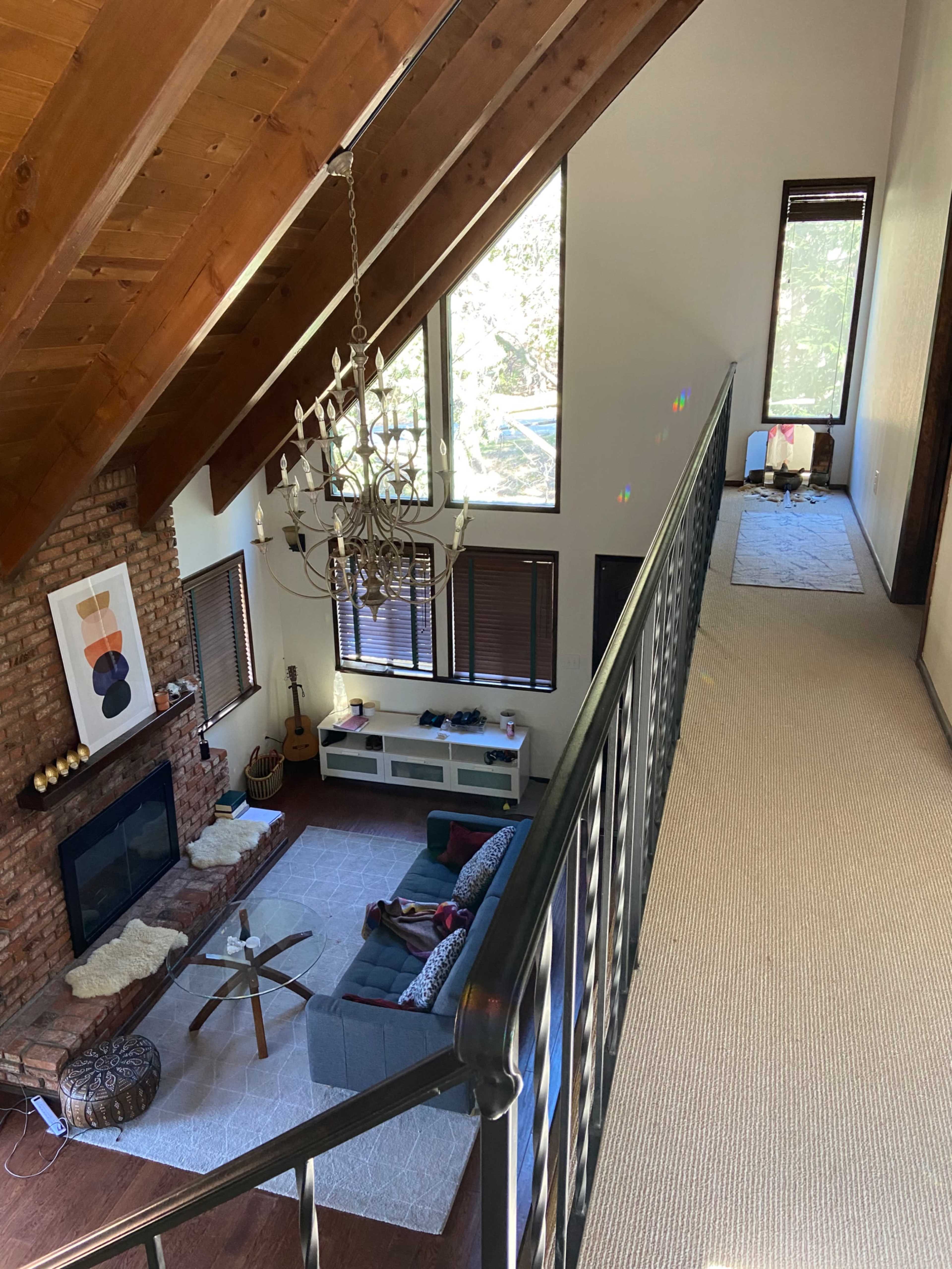 A spacious living room with a brick fireplace, a chandelier, and a view of a lofted area with a hallway and windows.