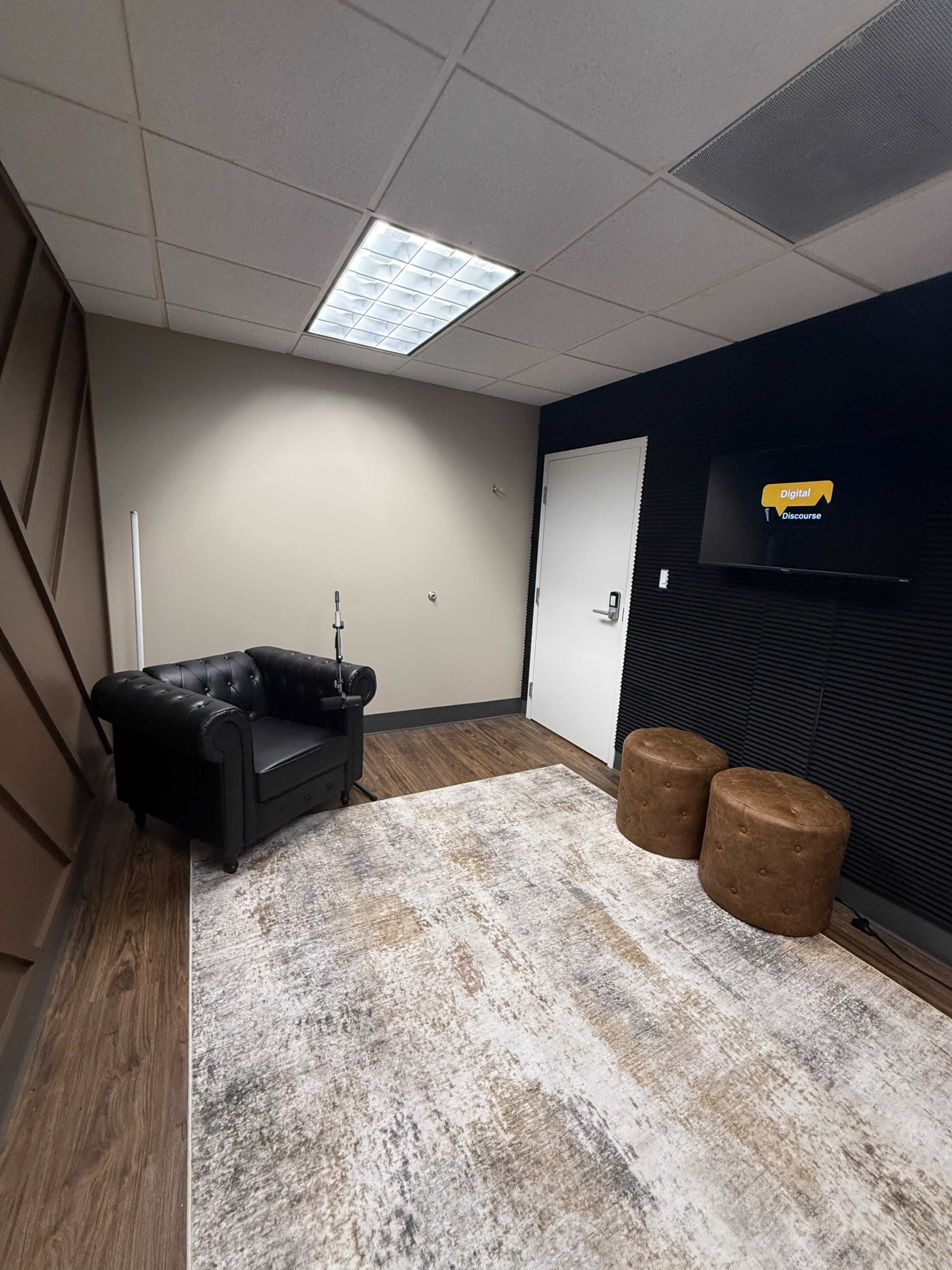 The image shows a small office space featuring a black leather chair, two brown stools, a patterned rug, and a television mounted on the wall.