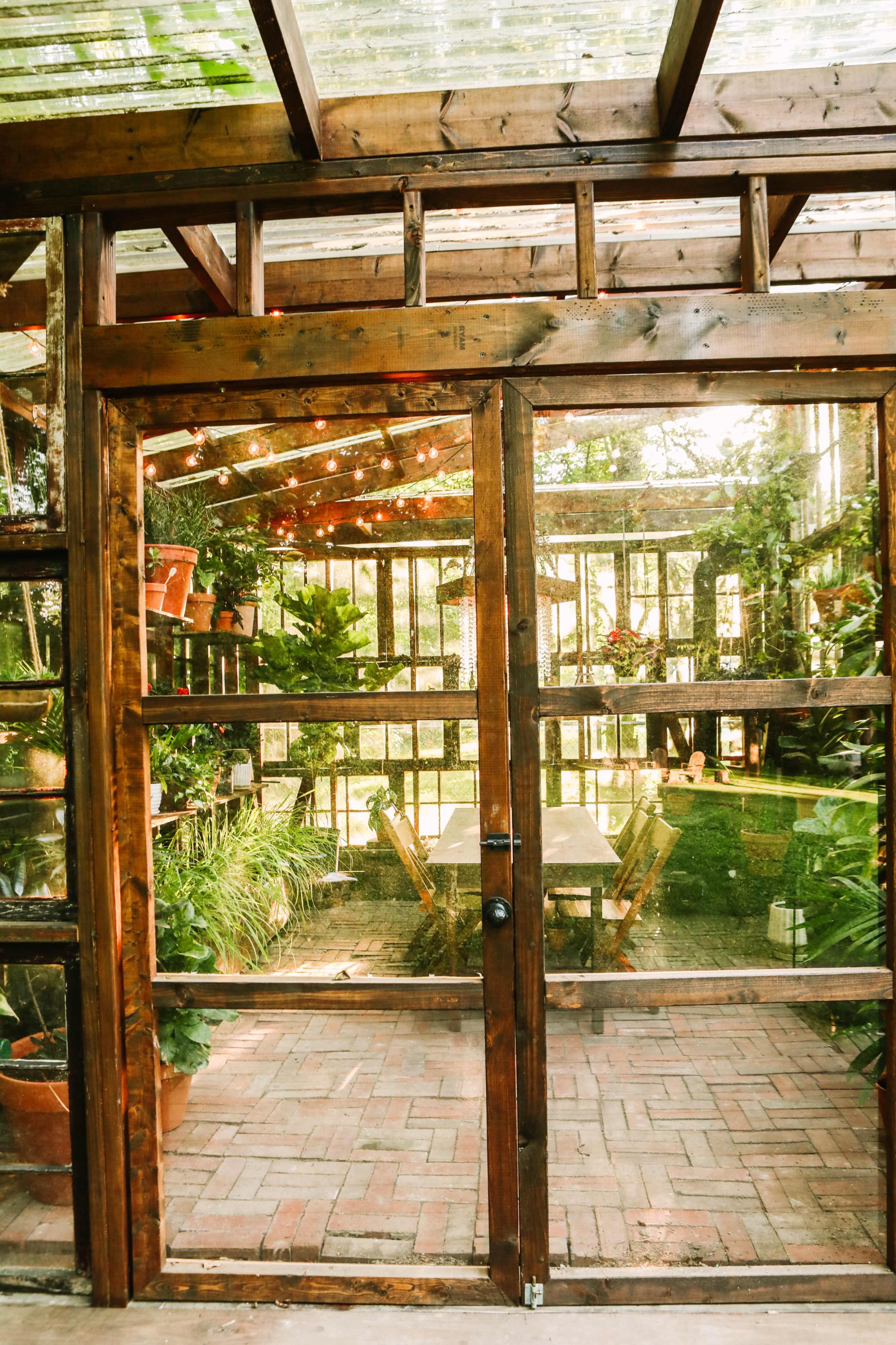 A wooden-framed greenhouse with a glass roof, filled with various plants and a set of tables inside, and a door that opens to the garden.