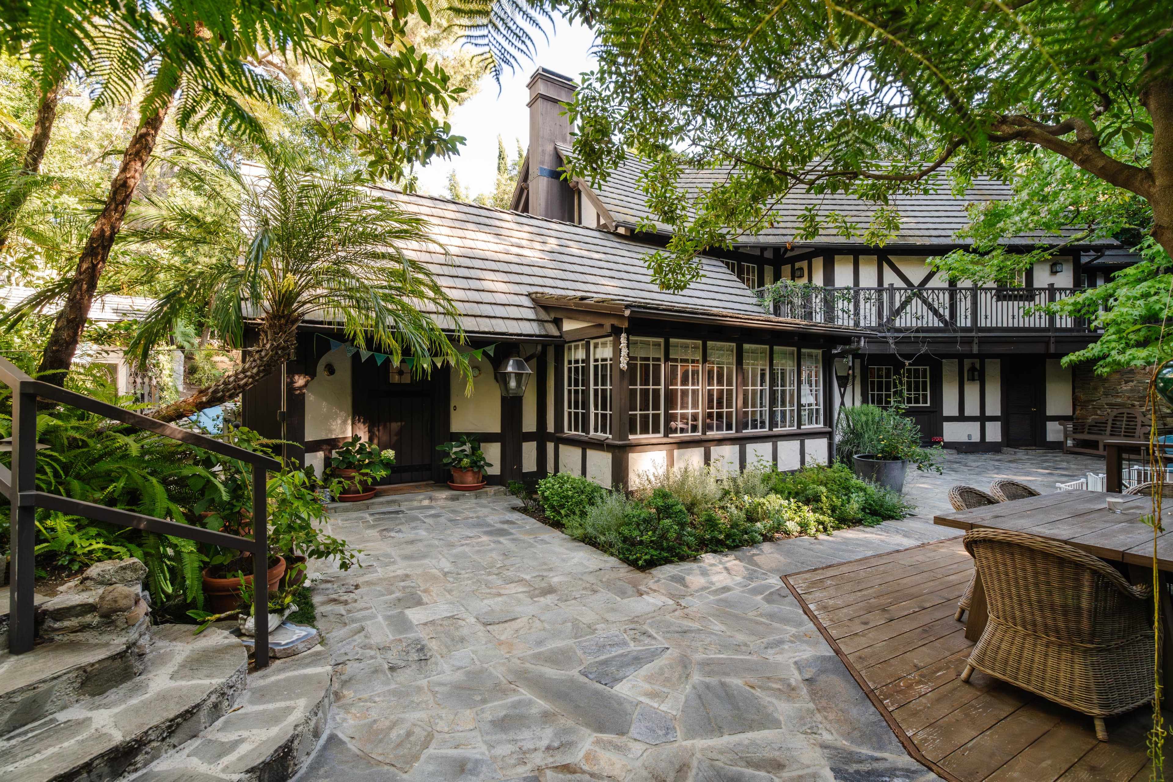 A stone pathway leading to a two-story, Tudor-style house surrounded by lush greenery and outdoor seating areas.