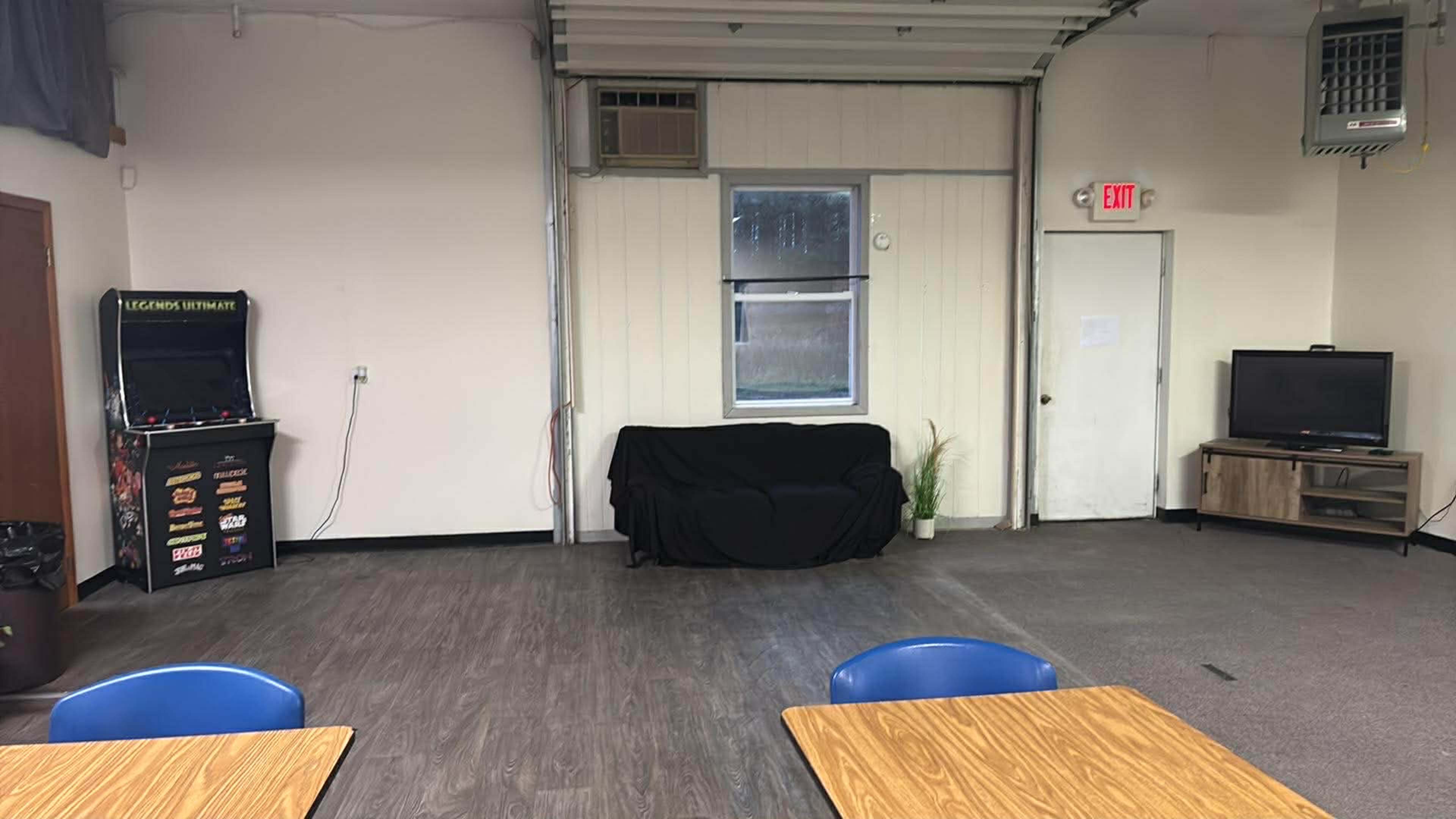 The image shows an indoor space with a black couch, a television, a window, a video game machine, and several tables with blue chairs.