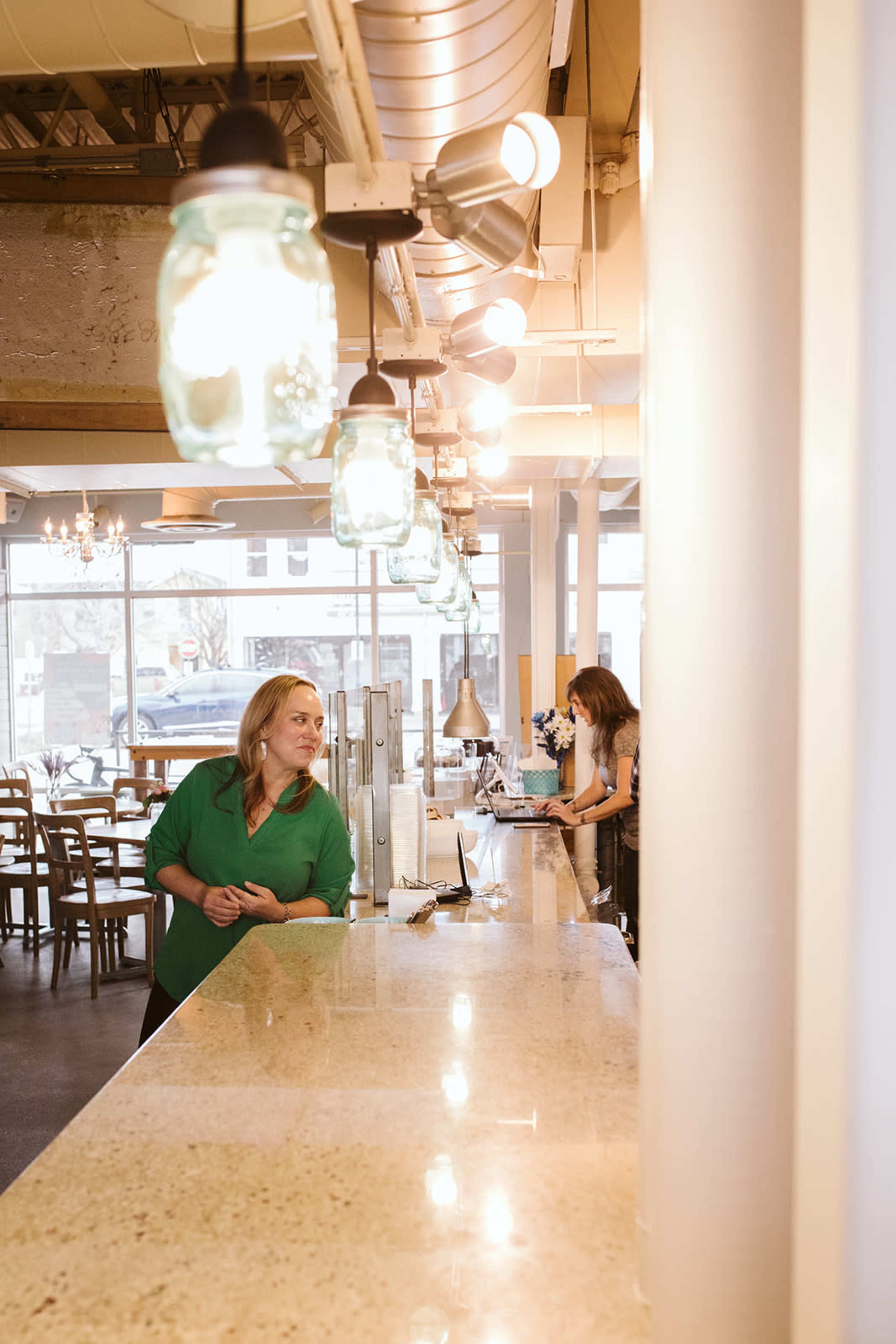 A woman stands at a counter while another woman works behind it in a bright café space with hanging lights.