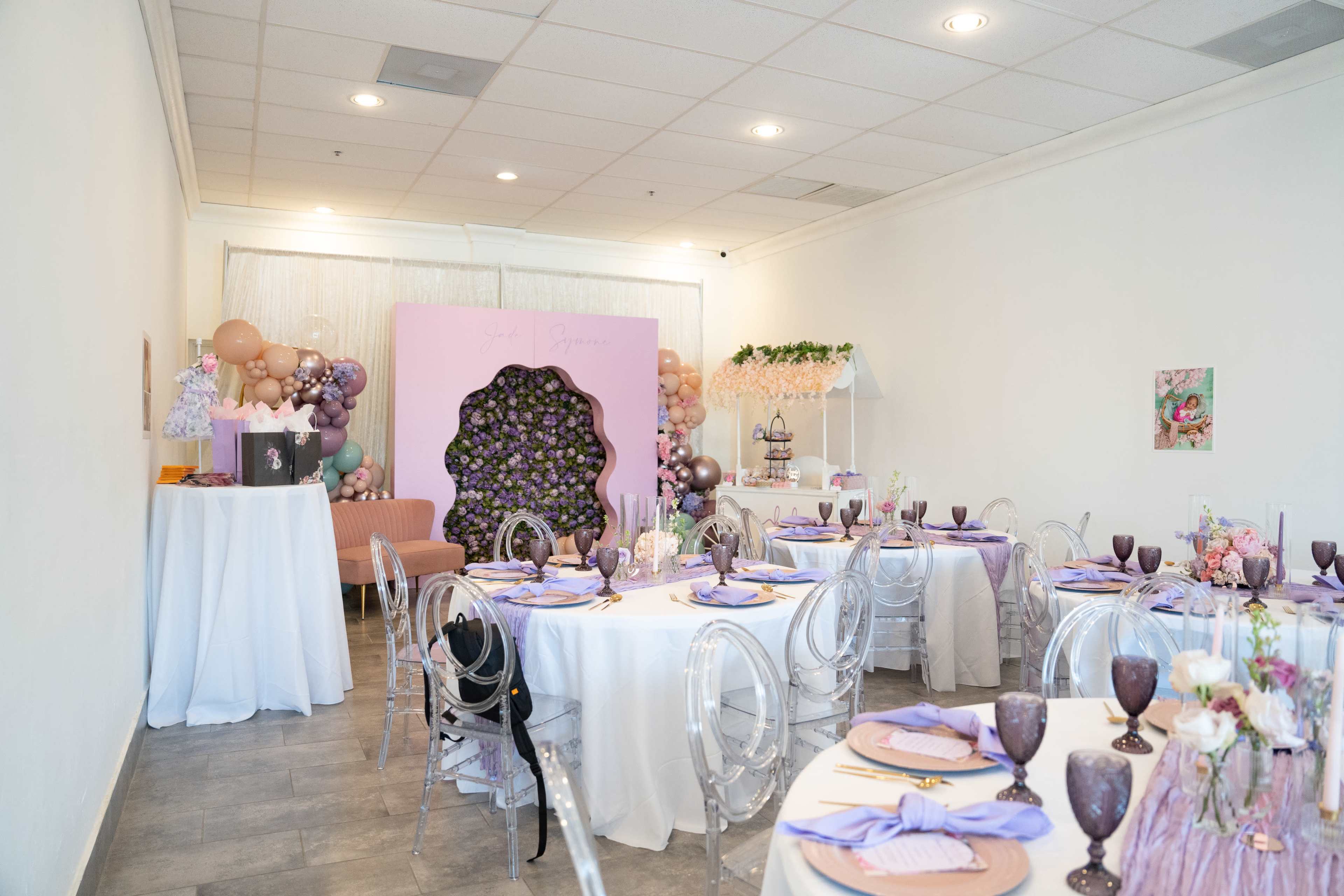 The image shows a decorated event space with tables set for a gathering, featuring purple accents, floral arrangements, and a decorative backdrop.