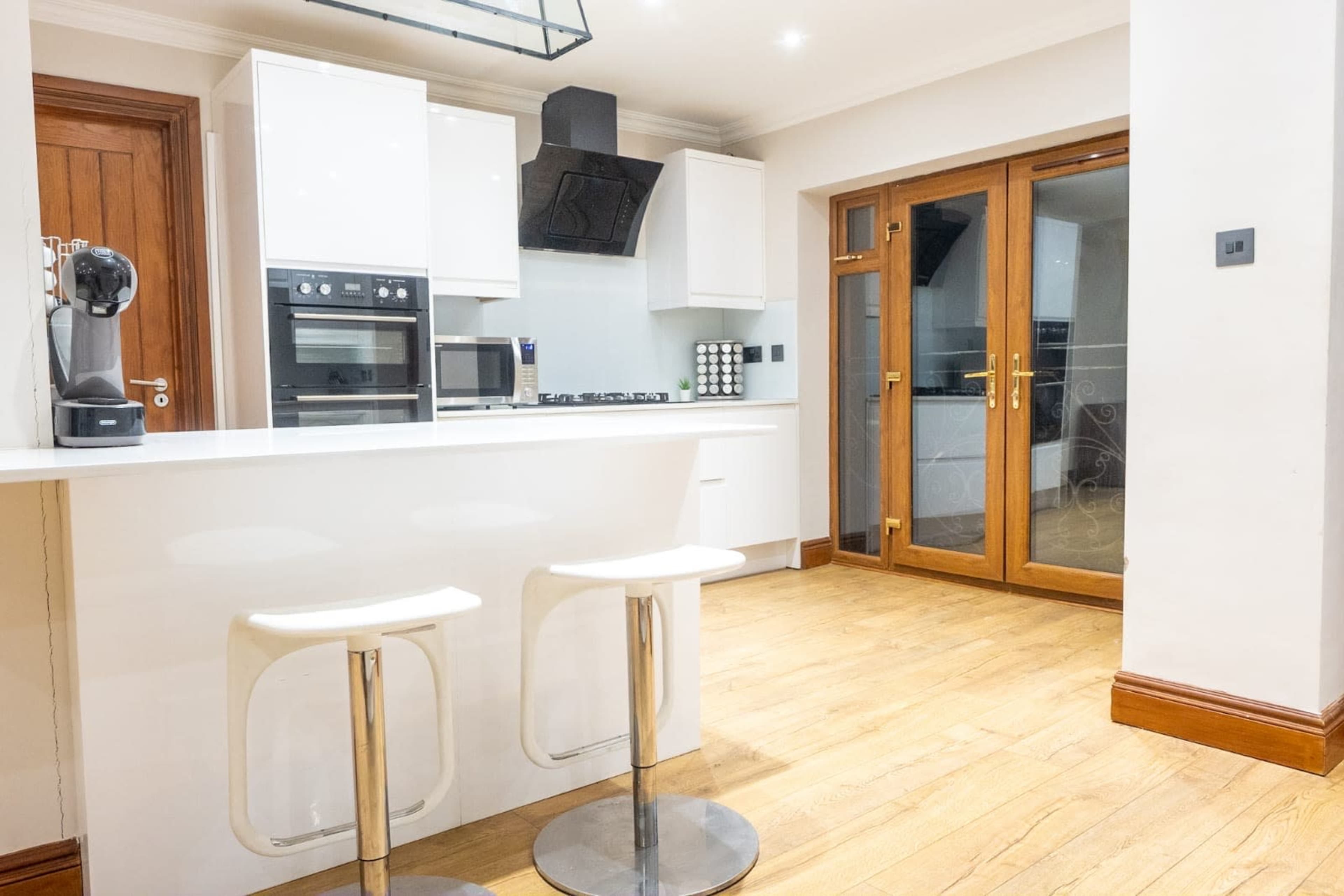 The image shows a modern kitchen with a white countertop, two stools, built-in appliances, and wooden flooring.