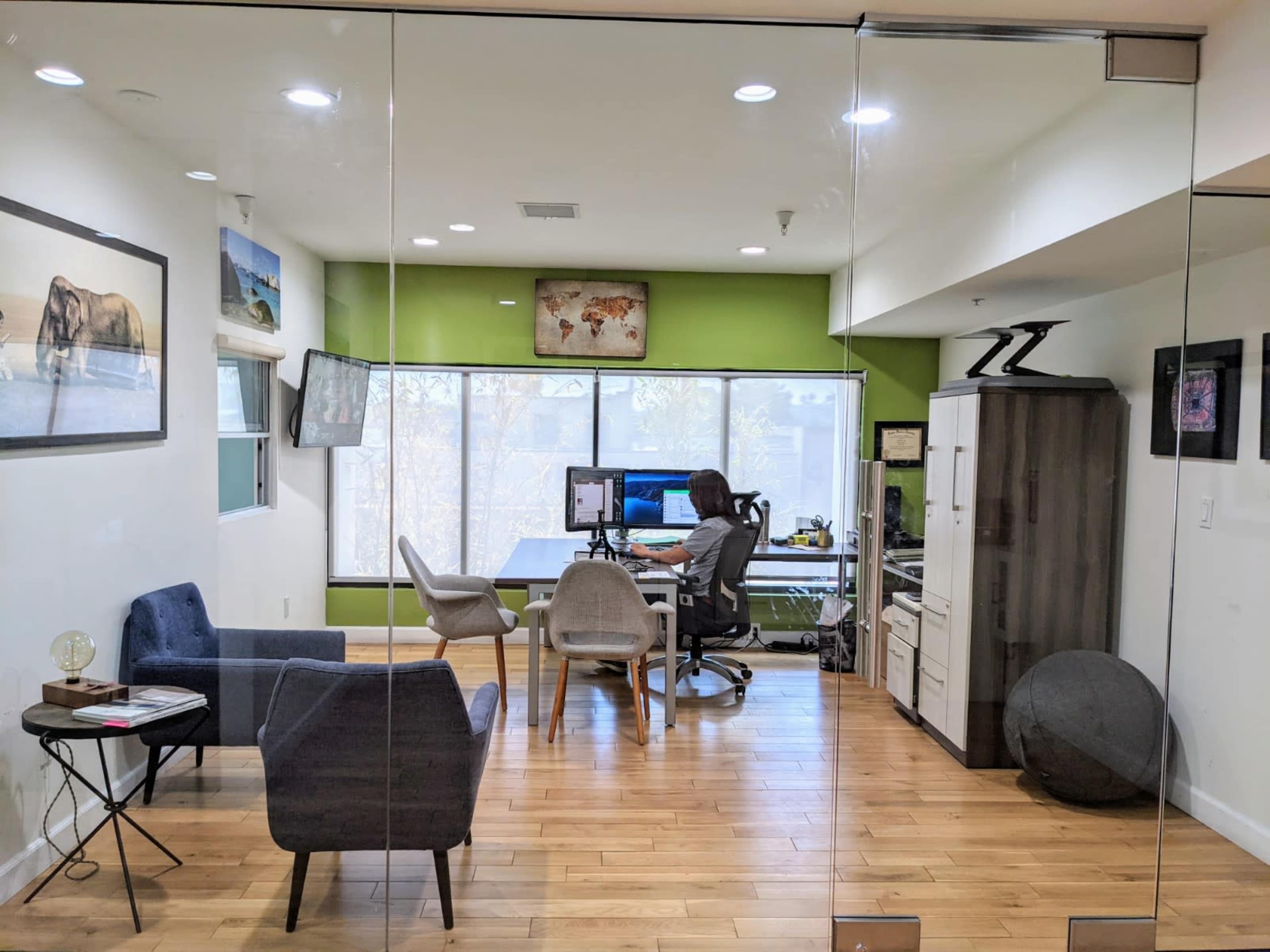 A person sits at a desk in a modern office with green walls, large windows, and various pieces of furniture.