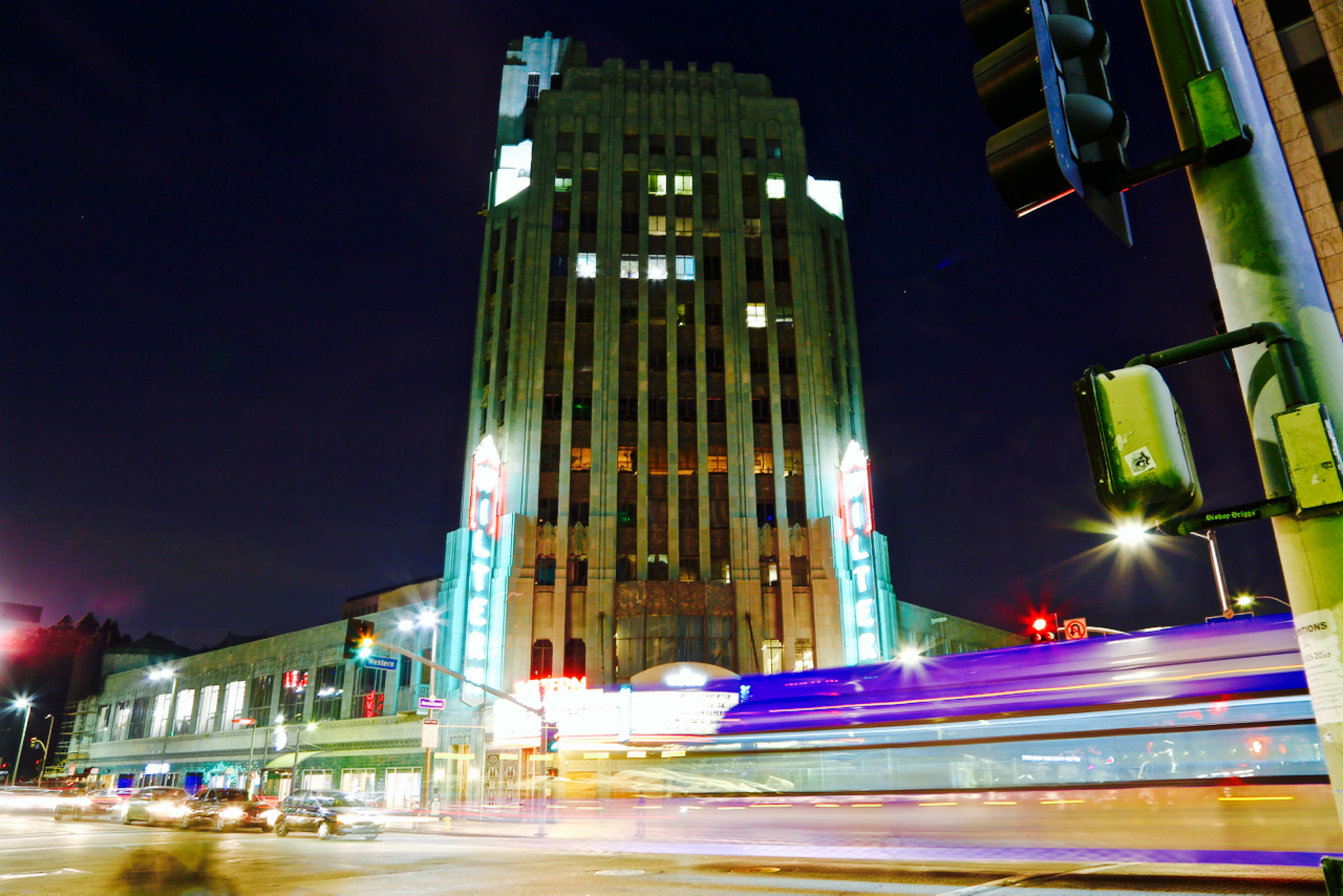 A tall, Art Deco building stands illuminated at night, with traffic flowing past and bright lights highlighting its facade.