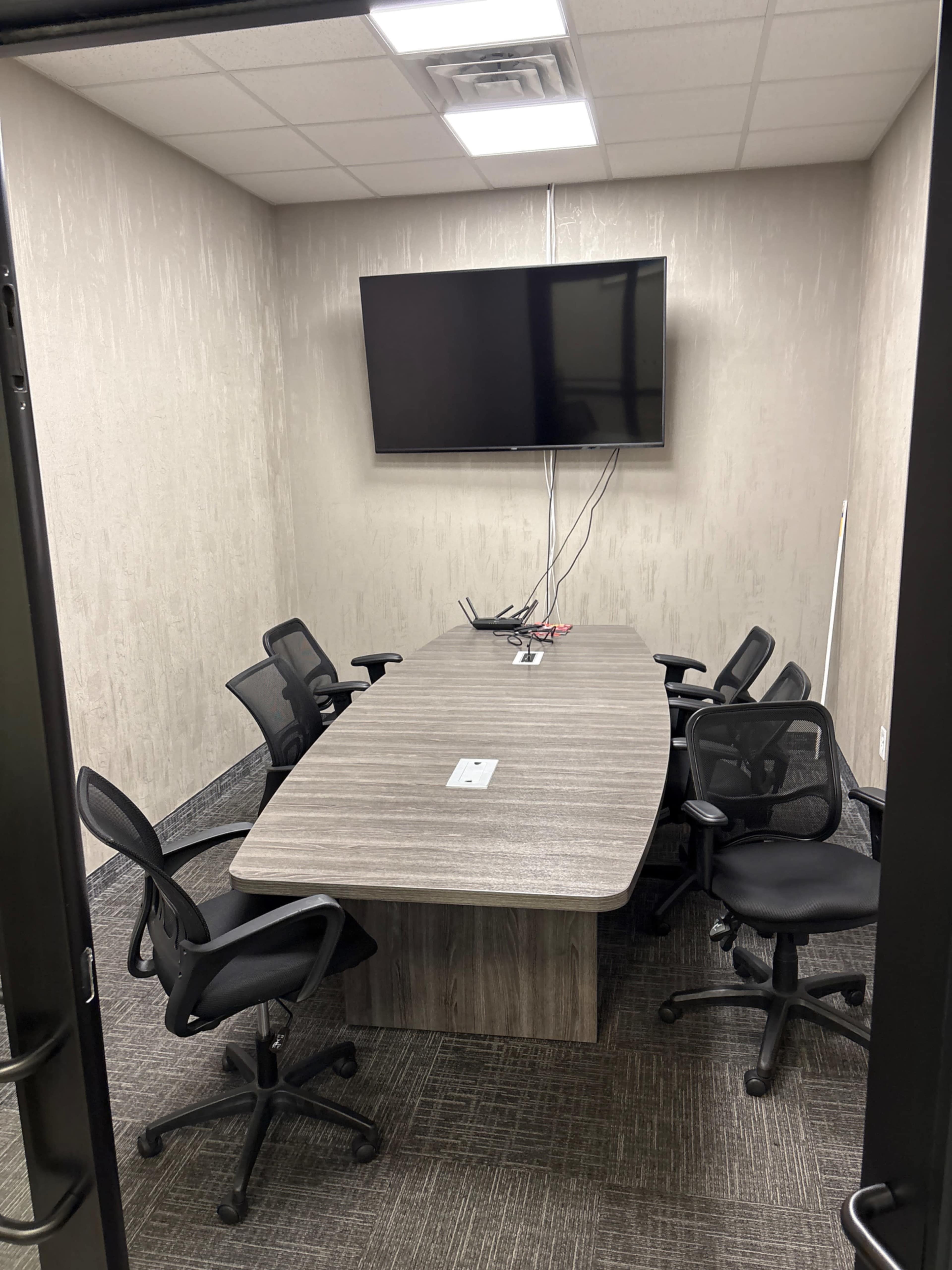 The image shows a meeting room with a long wooden table surrounded by six black office chairs and a wall-mounted television.