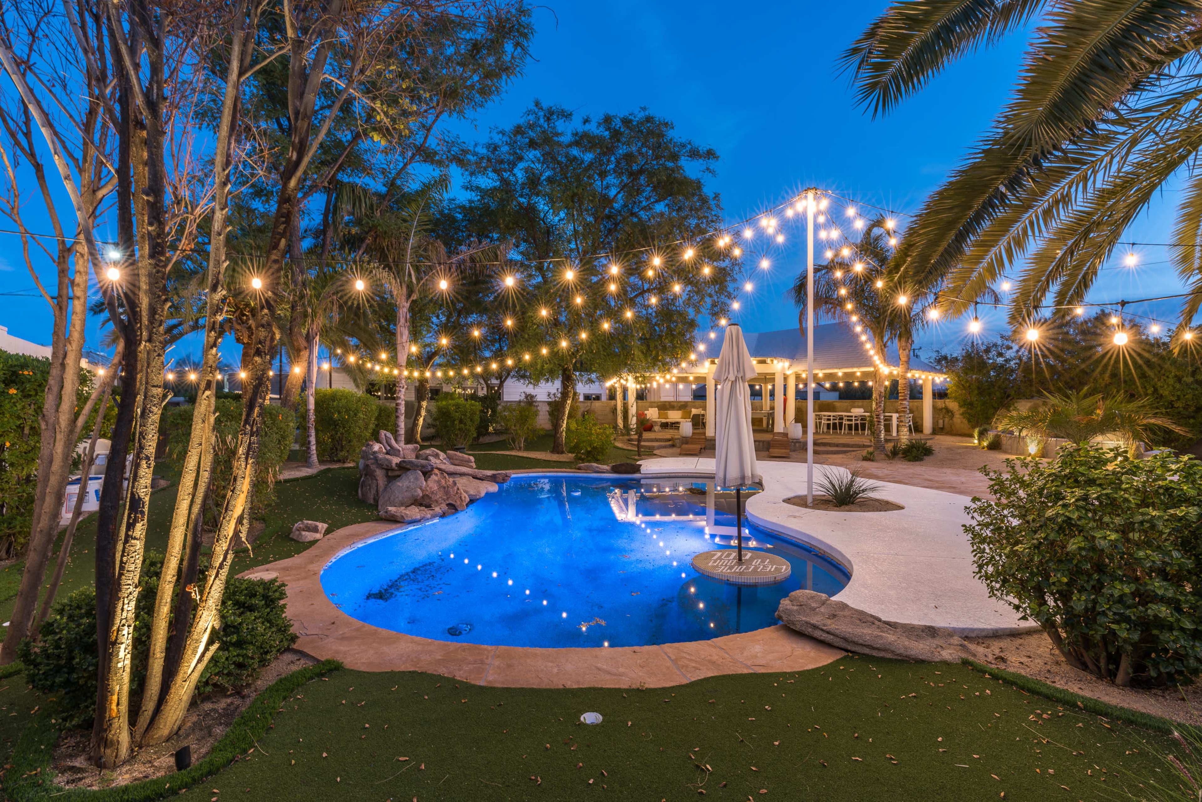 A backyard pool area with string lights above, surrounded by landscaped greenery and a shaded patio.