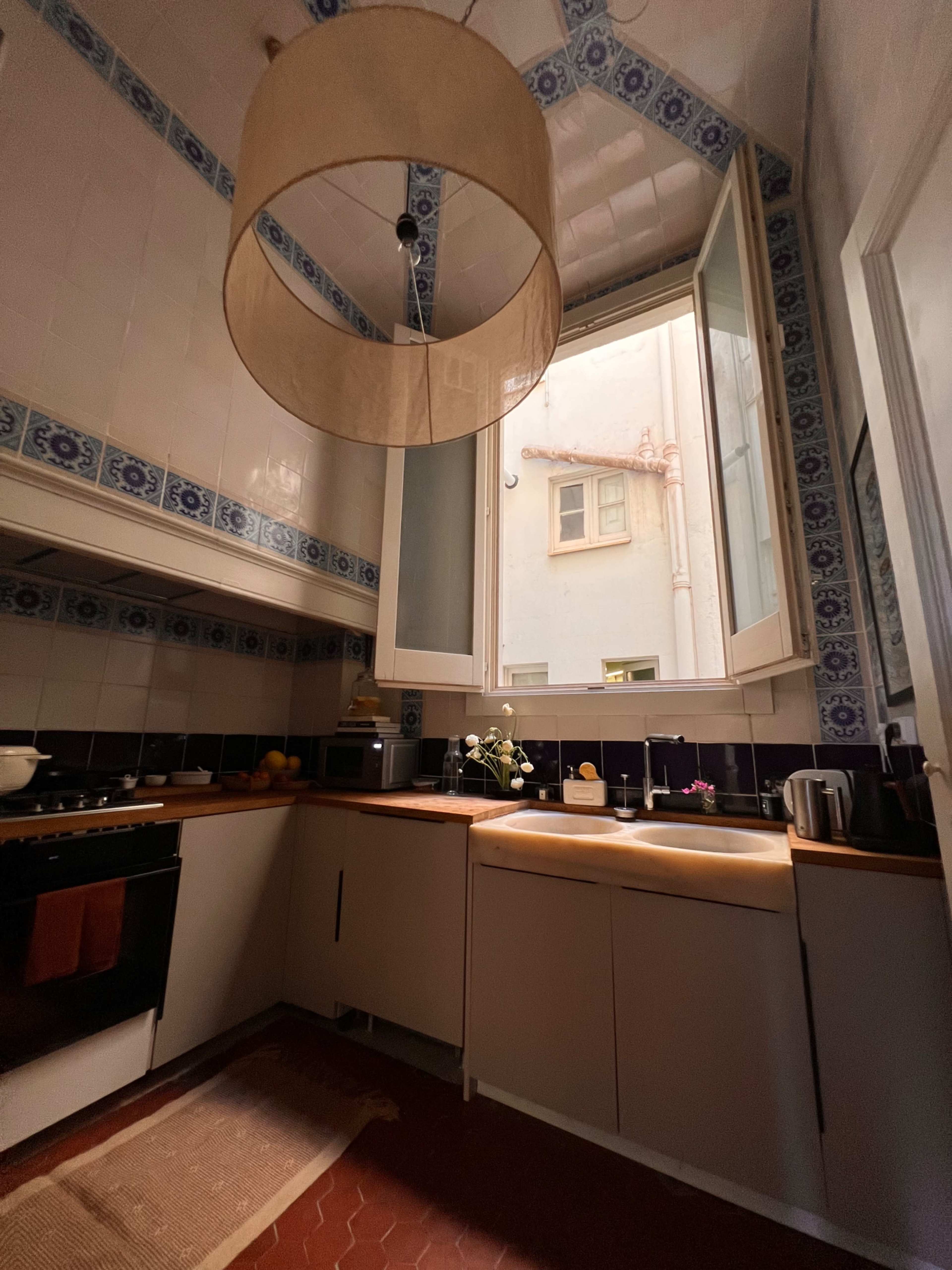 A small kitchen features wooden countertops, a ceiling lamp, and two open windows overlooking an exterior courtyard.