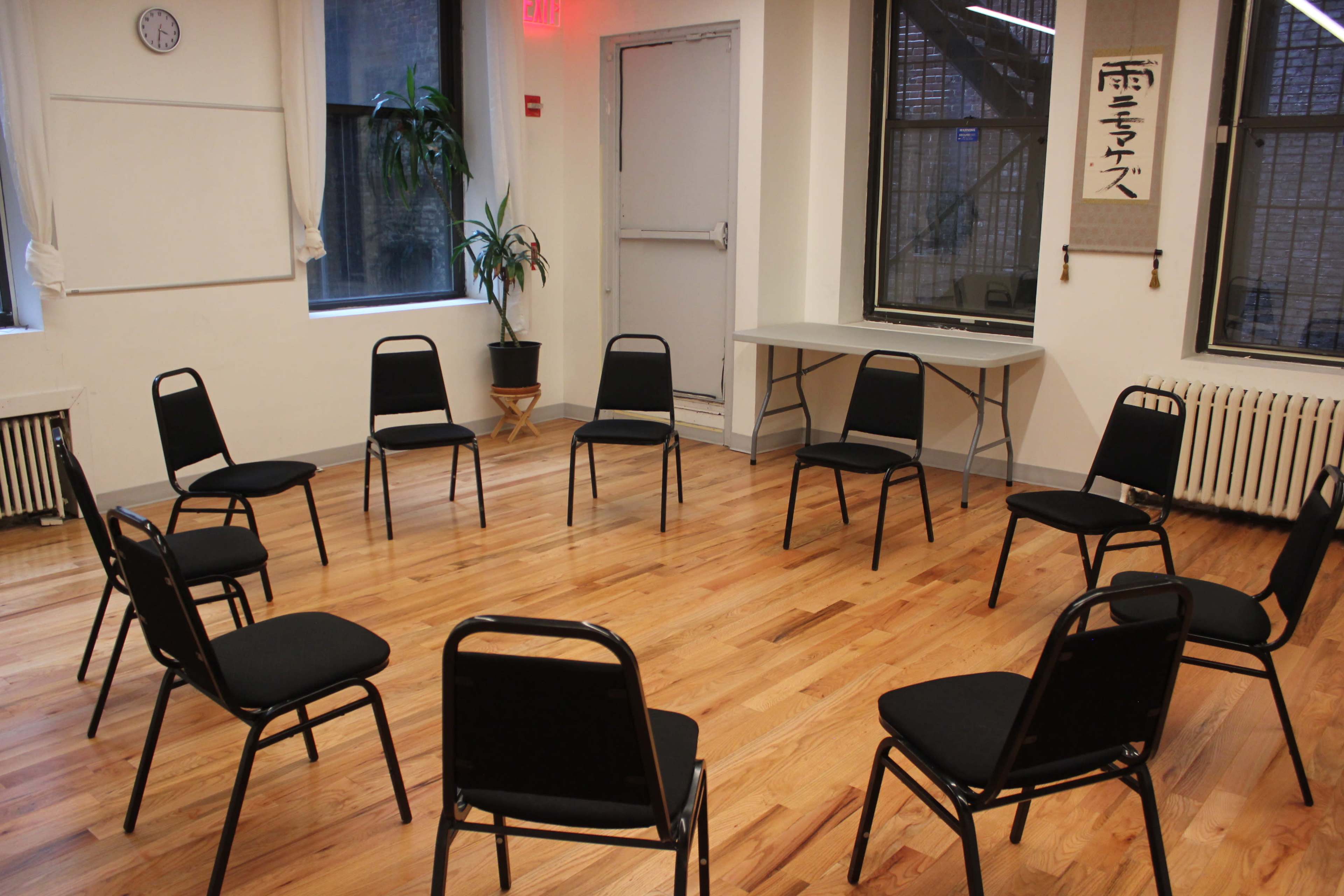 A circle of black chairs is arranged on a wooden floor in a well-lit room with large windows and a potted plant.