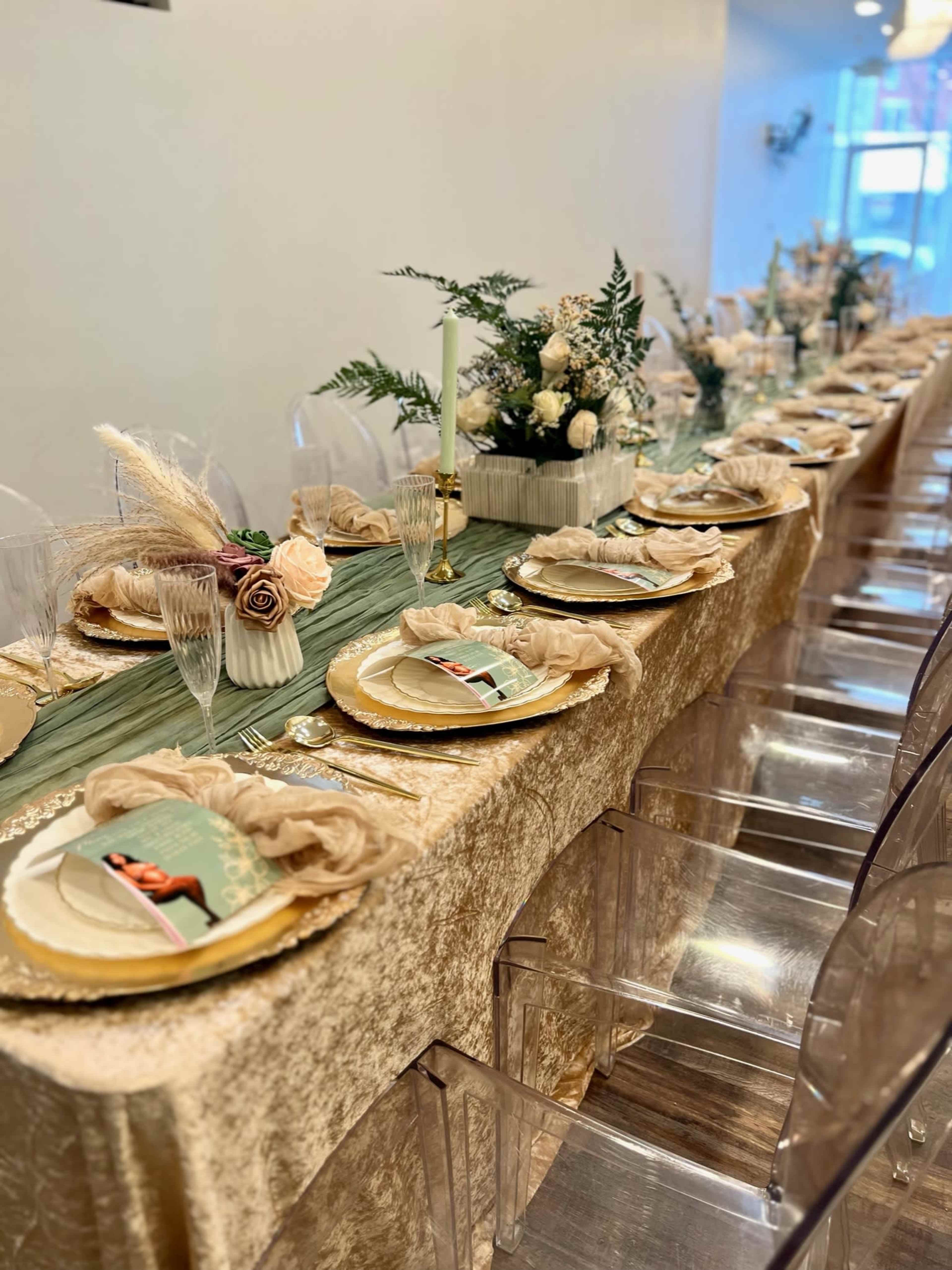 A elegantly set dining table with a gold tablecloth, clear chairs, plates, decorative flowers, and styled place settings.