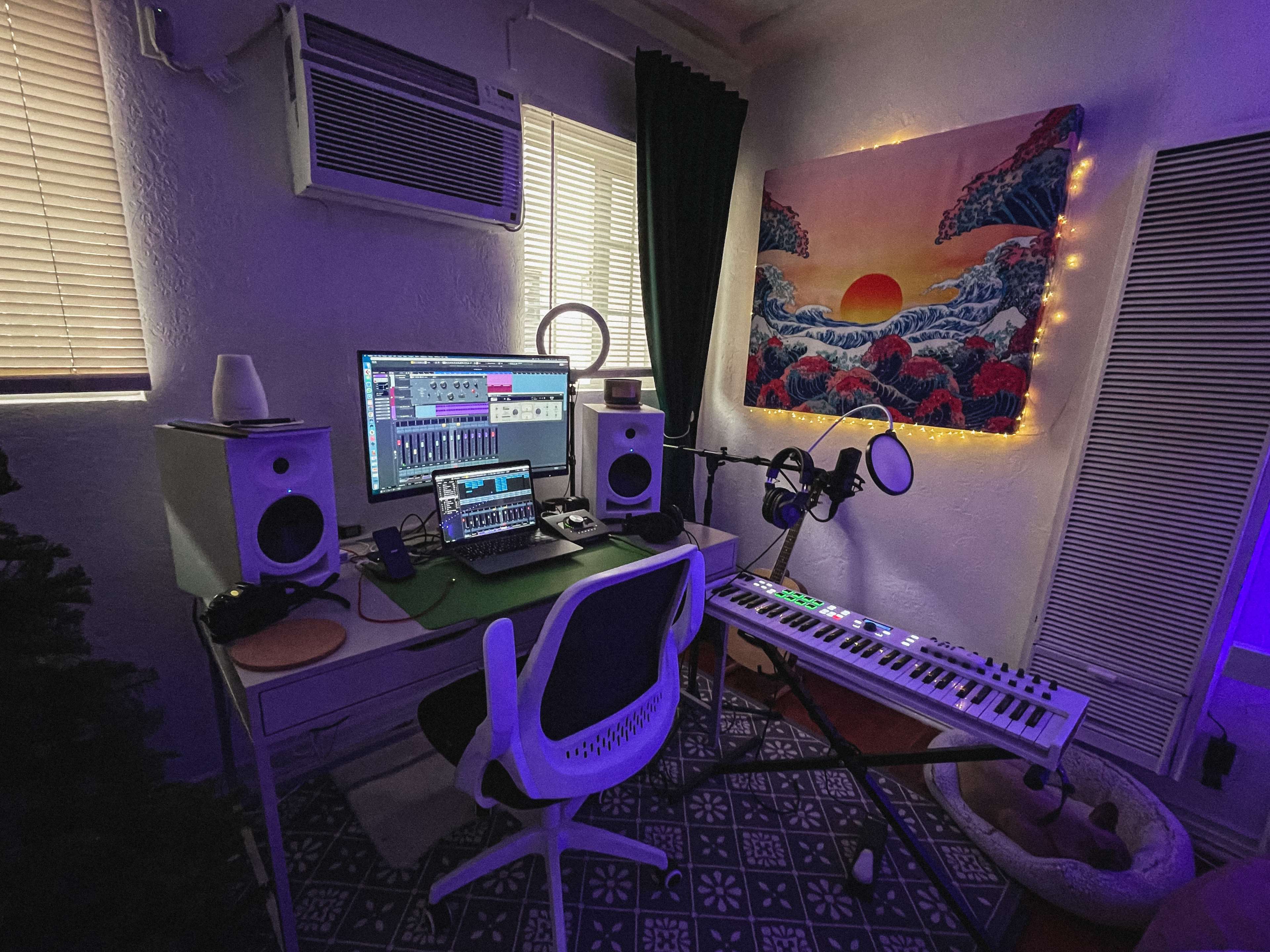 A home music studio features a desk with computer monitors, audio equipment, a MIDI keyboard, and decorative lighting on the walls.