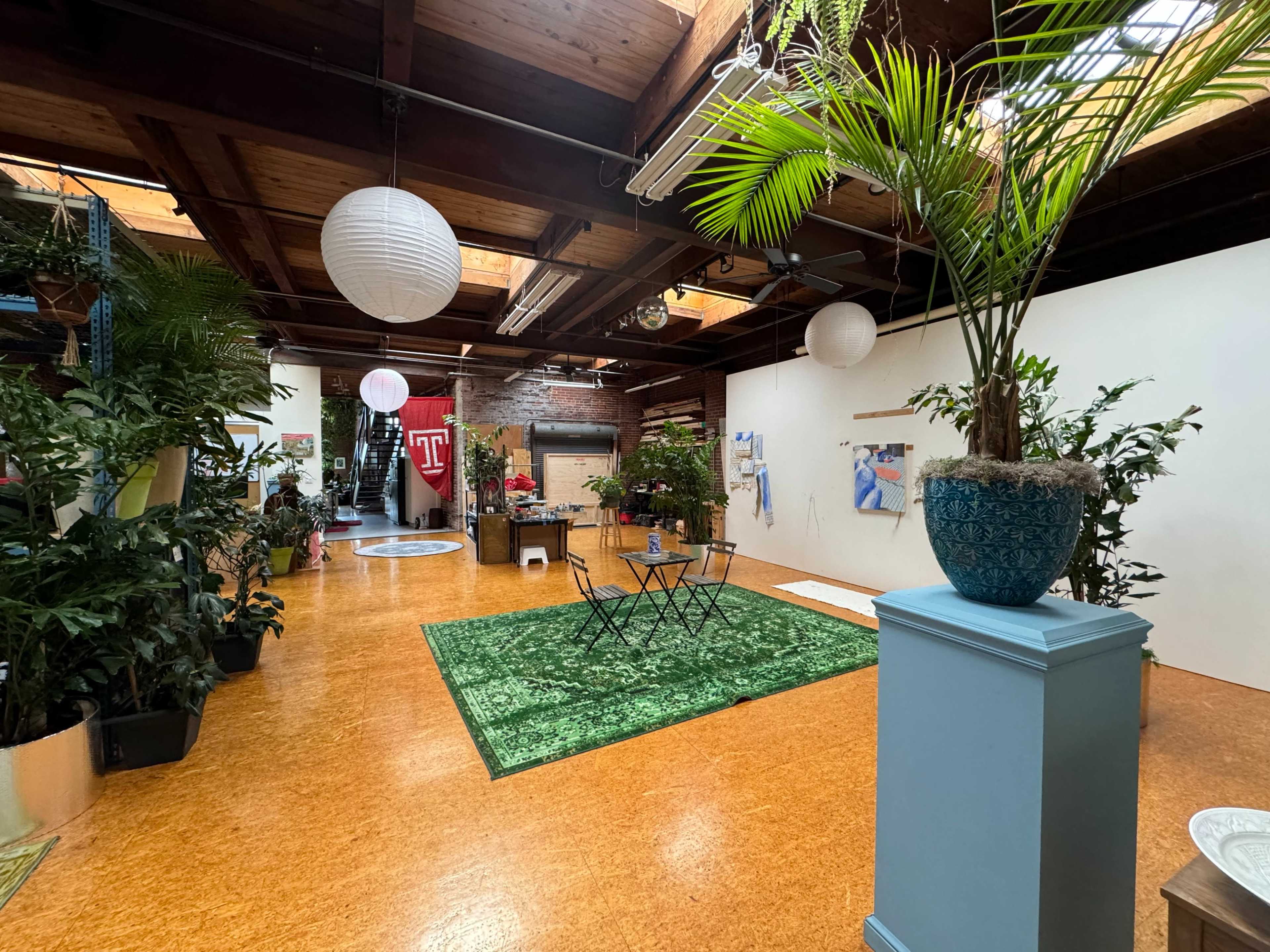 A spacious interior with wooden beams and a variety of indoor plants, featuring a green carpet, a small table with chairs, and decorative lighting.