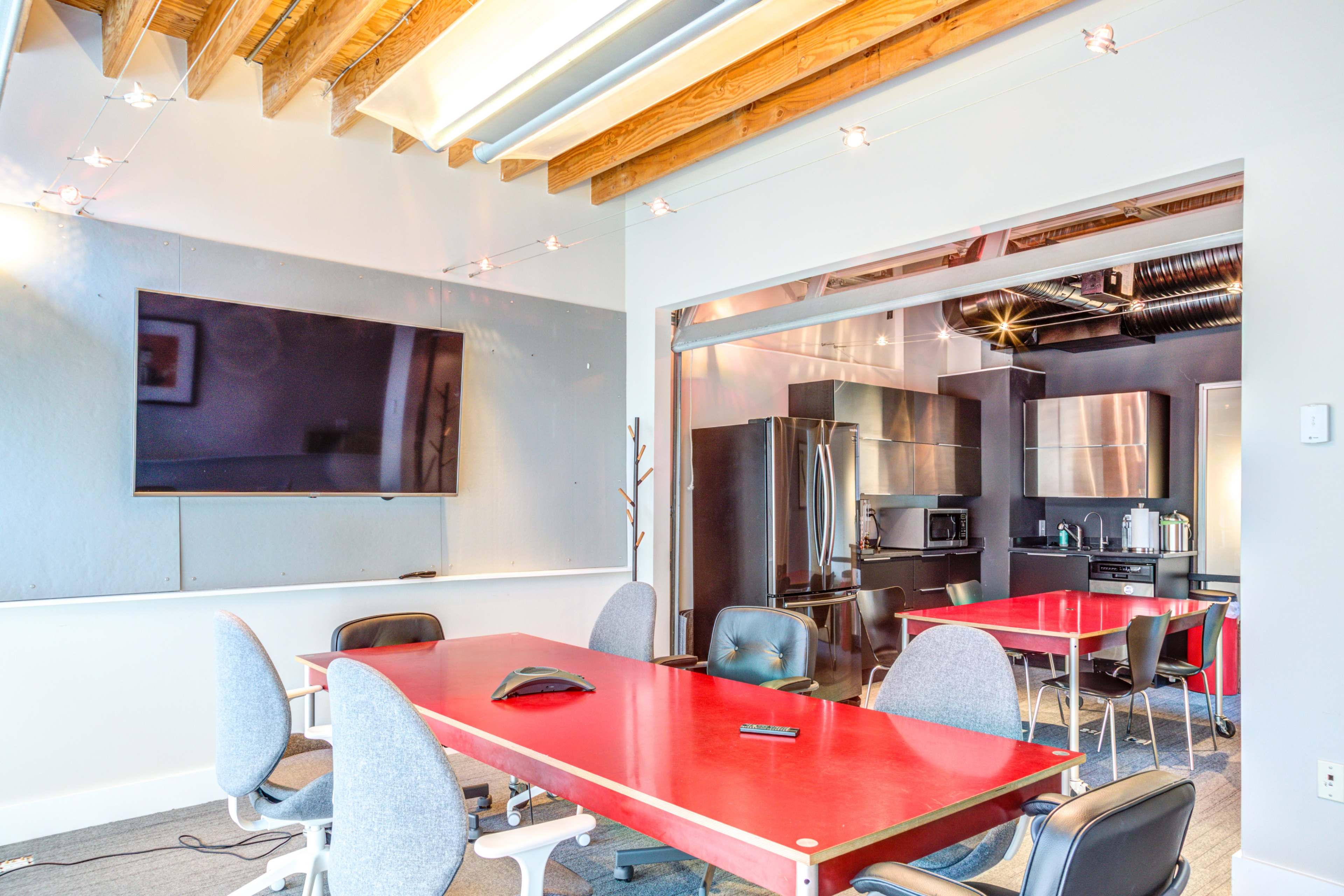 A modern conference room features a red table, ergonomic chairs, and a large wall-mounted screen, with a kitchenette visible in the background.