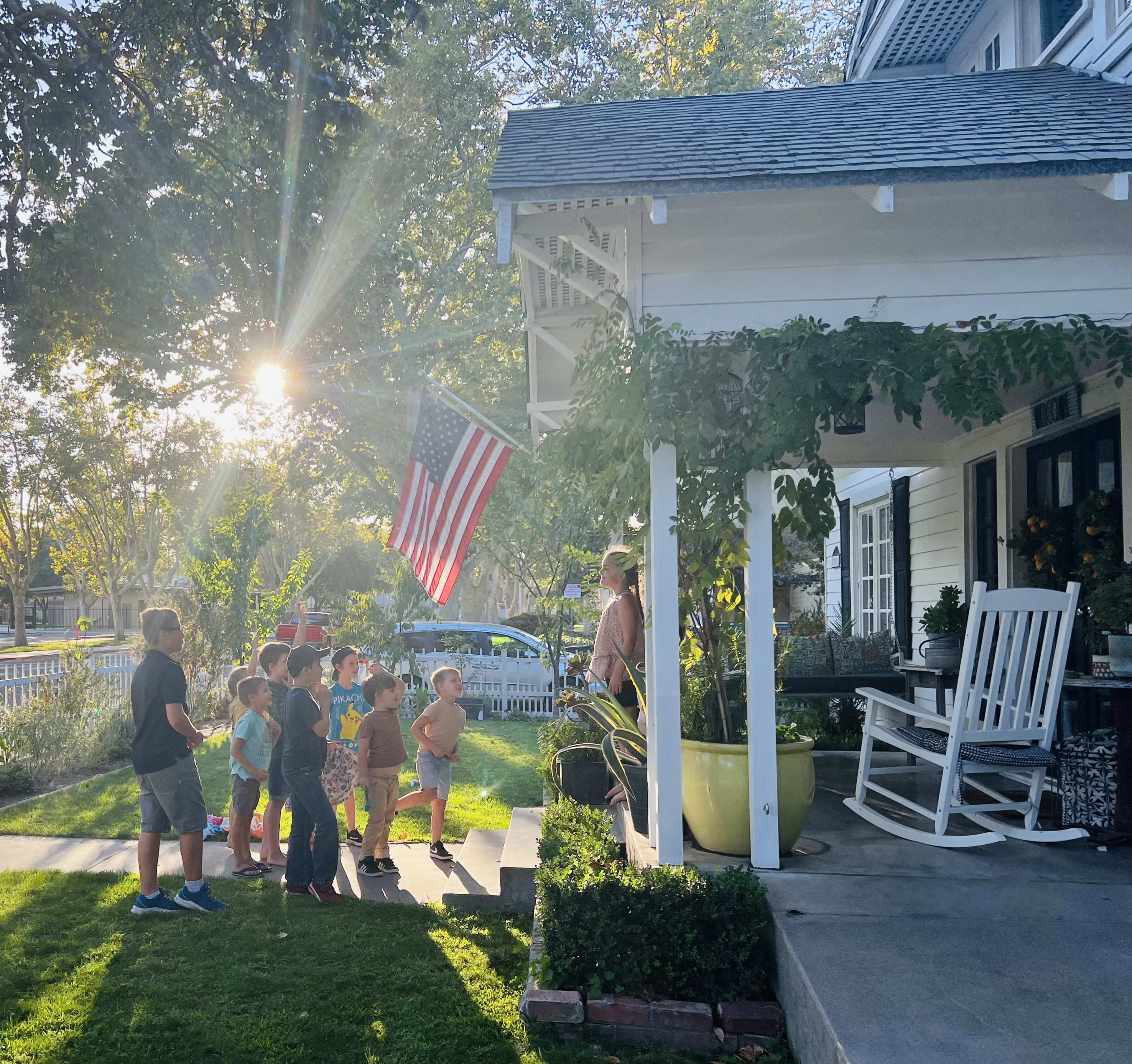 A group of children stands in a line on the lawn in front of a house, with a parent speaking to them under a porch adorned with an American flag.