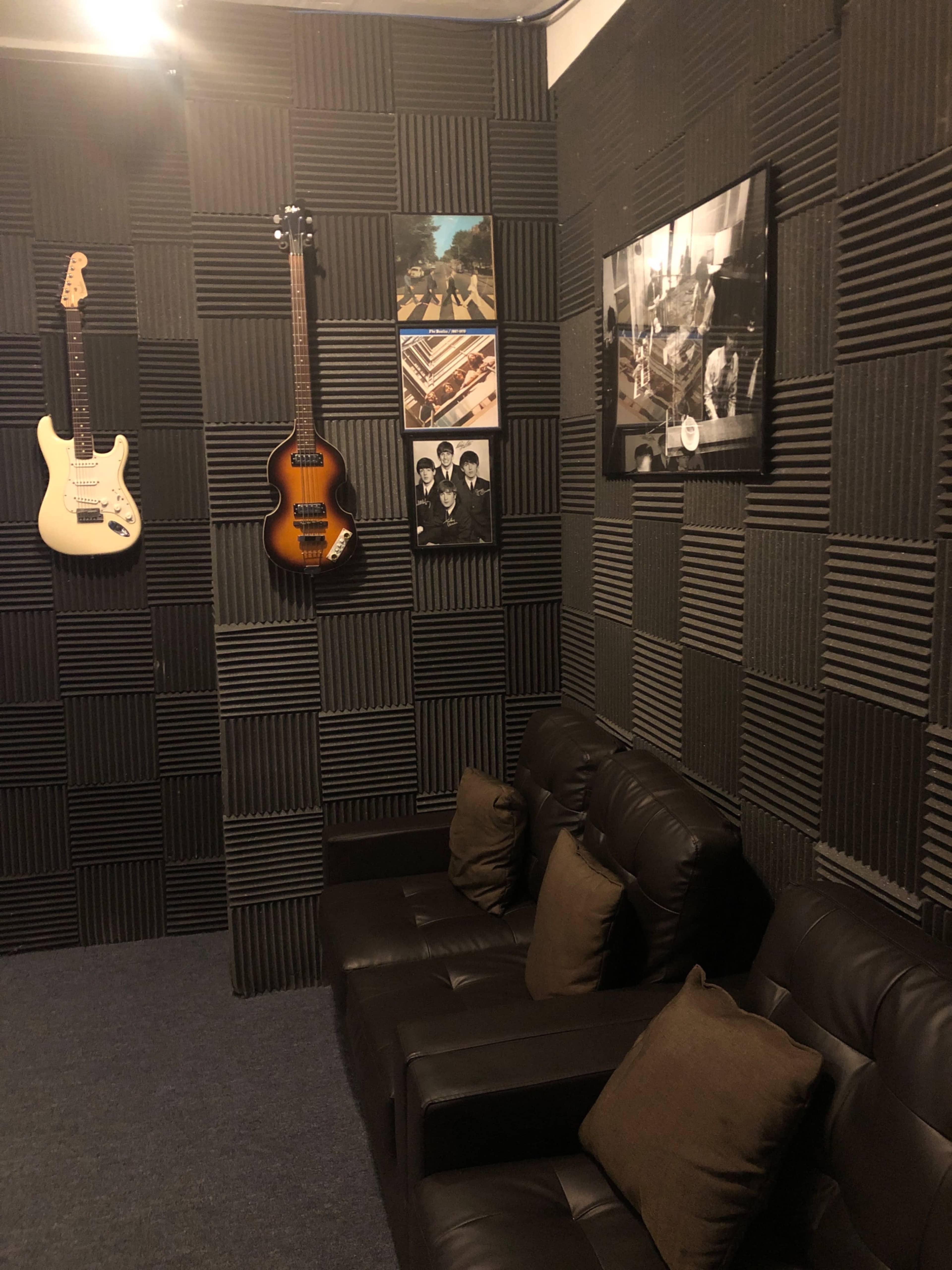 A soundproofed room featuring two guitars mounted on the wall, framed pictures, and a dark leather sofa with cushions.