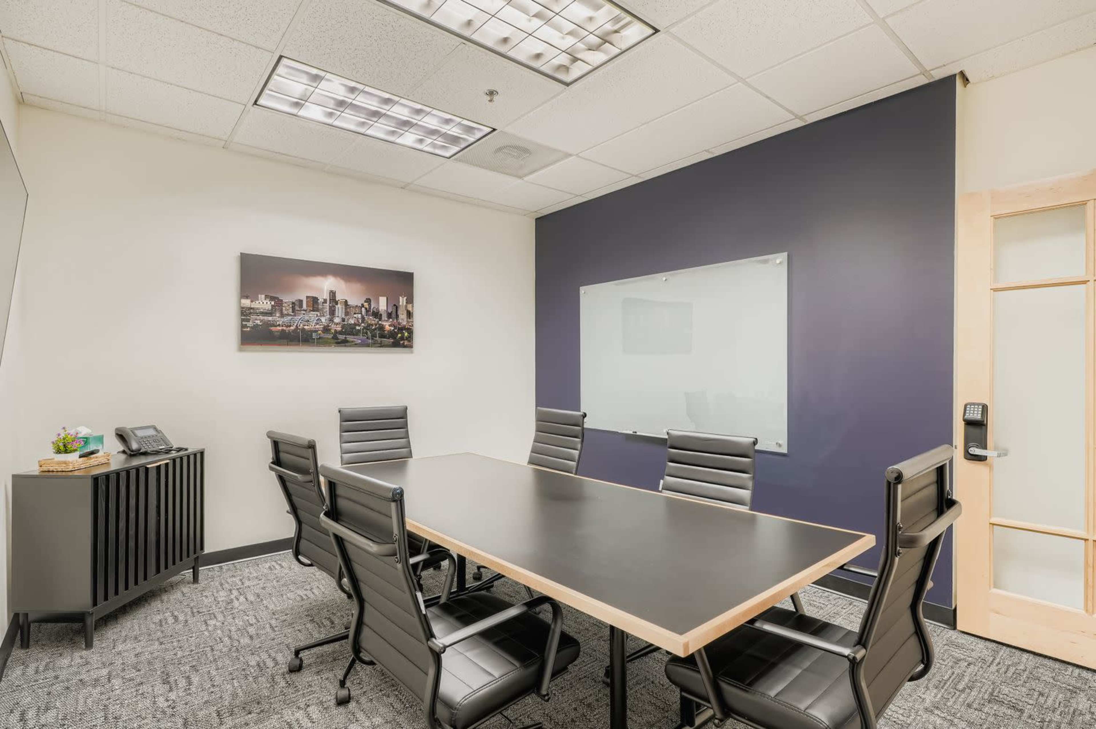 The image shows a conference room with a rectangular table surrounded by ergonomic black chairs, a whiteboard, and a framed cityscape photo on the wall.