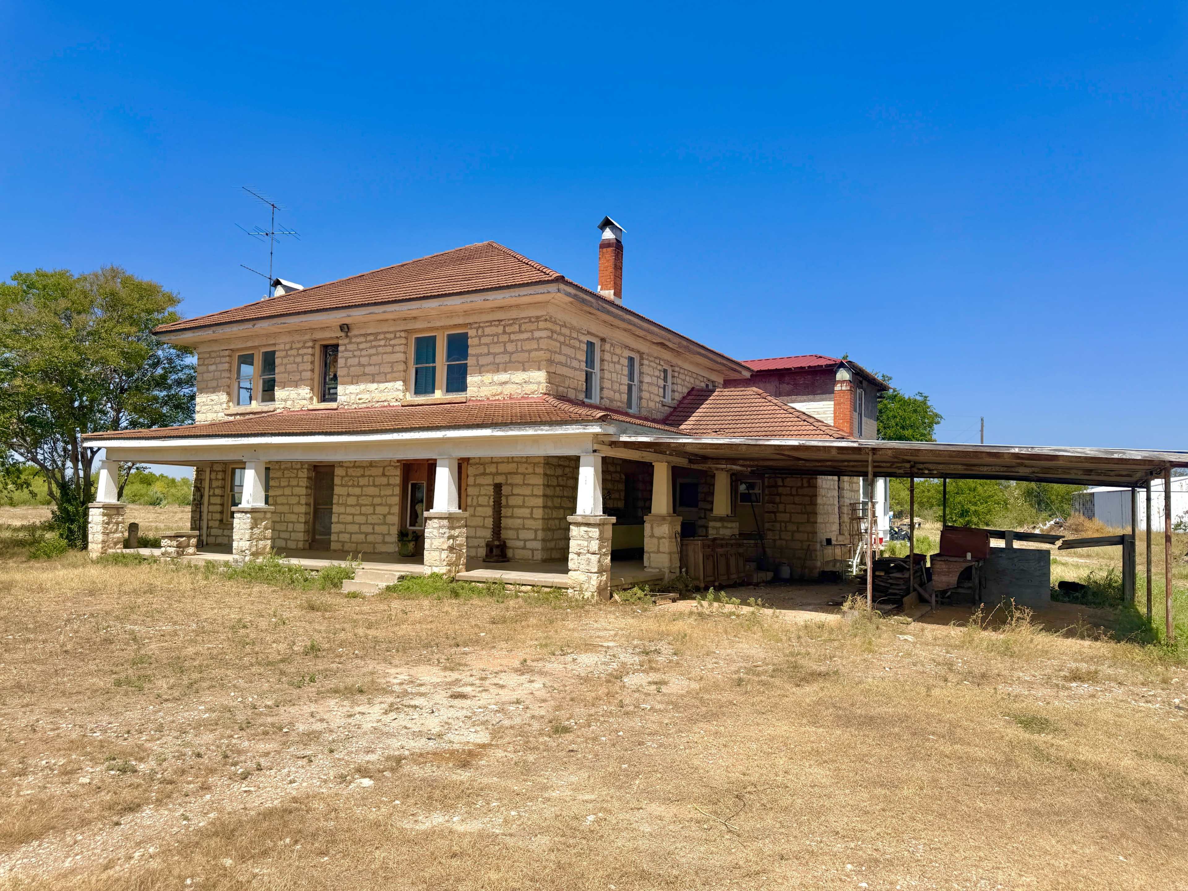 A two-story stone house with a red roof and covered porch sits on a grassy area against a clear blue sky.