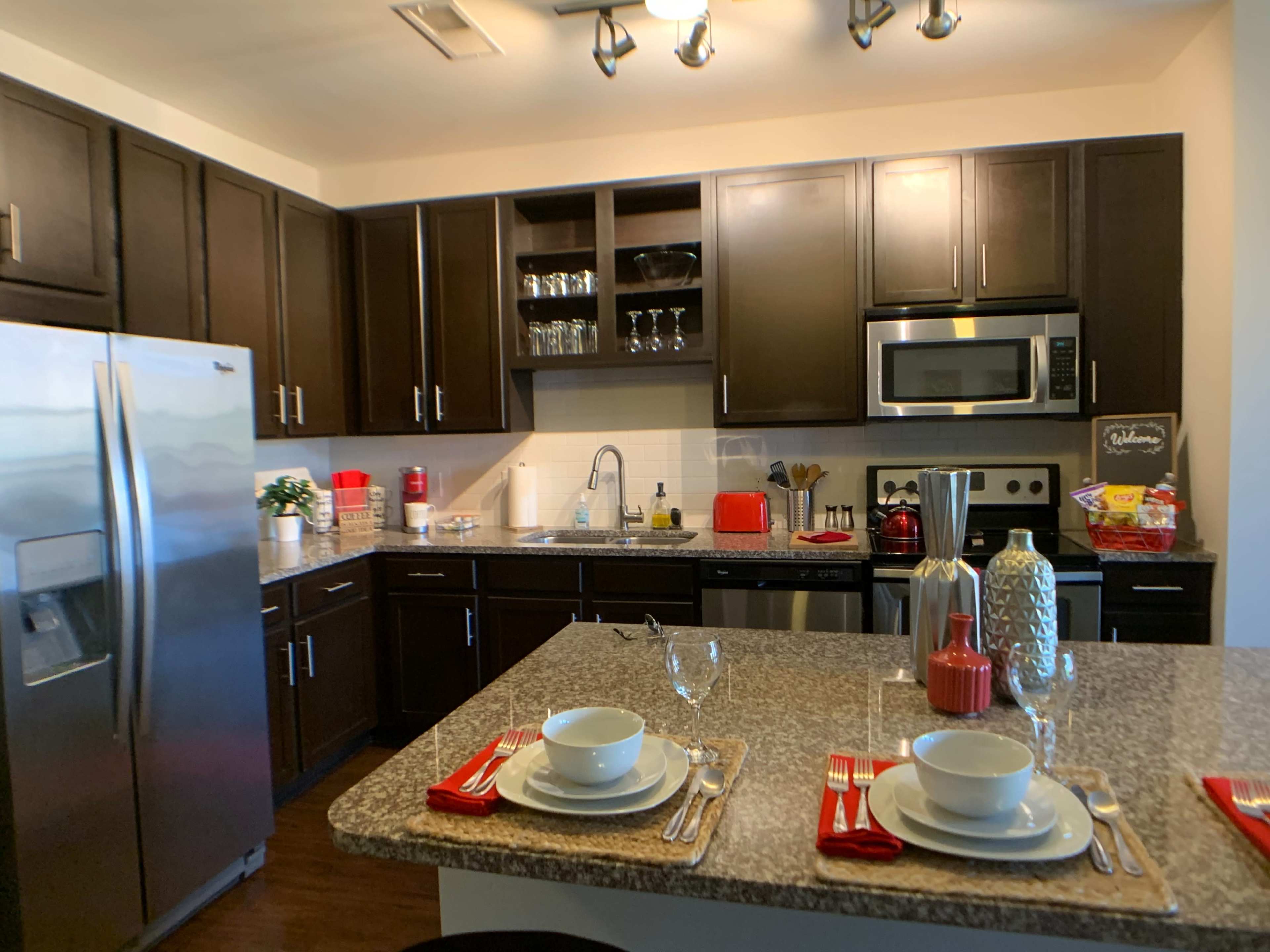 A modern kitchen with dark cabinetry, a stainless steel refrigerator, and a countertop set for two with plates and wine glasses.