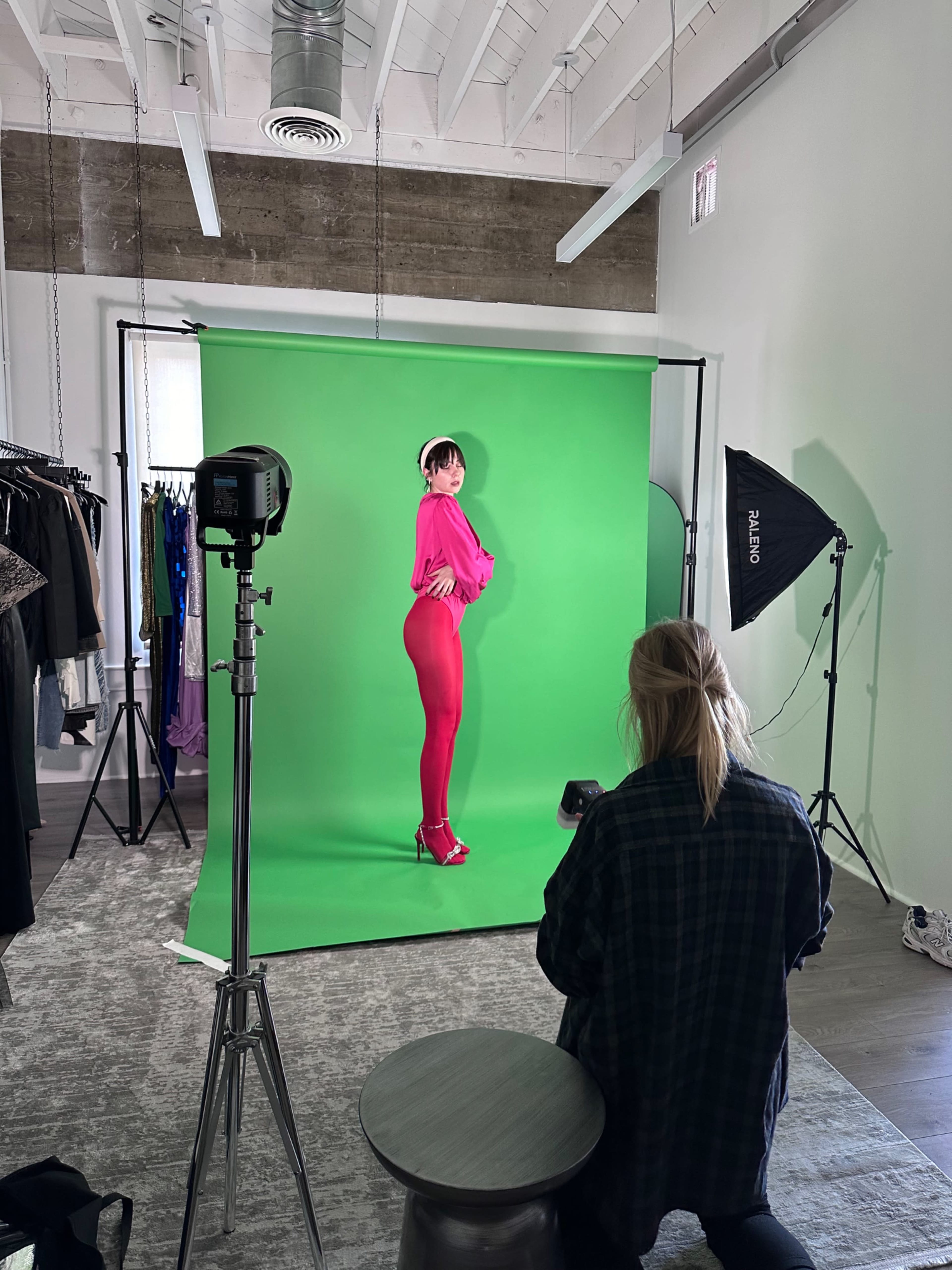 A model poses in a bright pink outfit against a green backdrop while a photographer captures the shot.