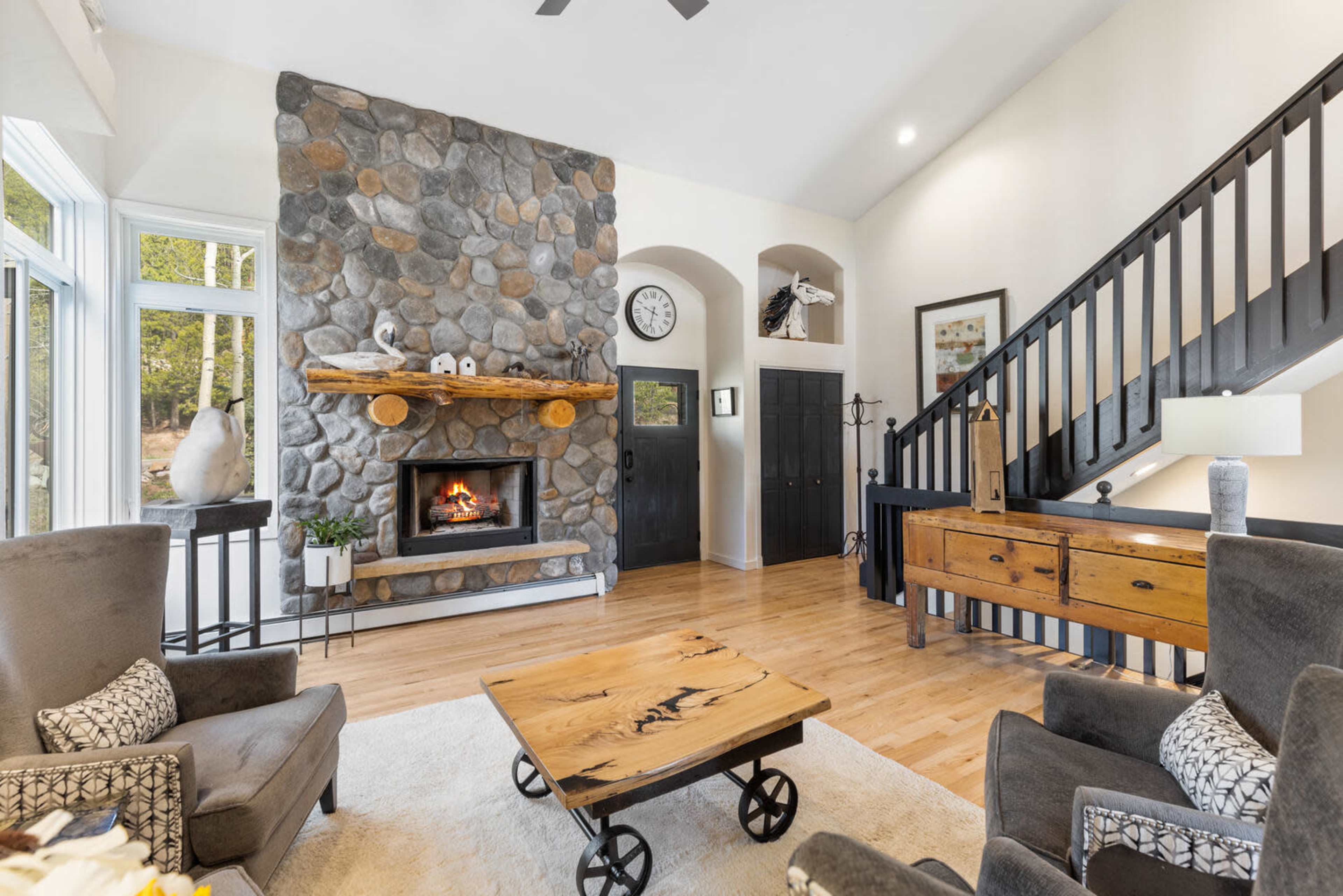 The image depicts a cozy living room featuring a stone fireplace, wooden mantel, and a staircase, with comfortable seating and a coffee table in the center.