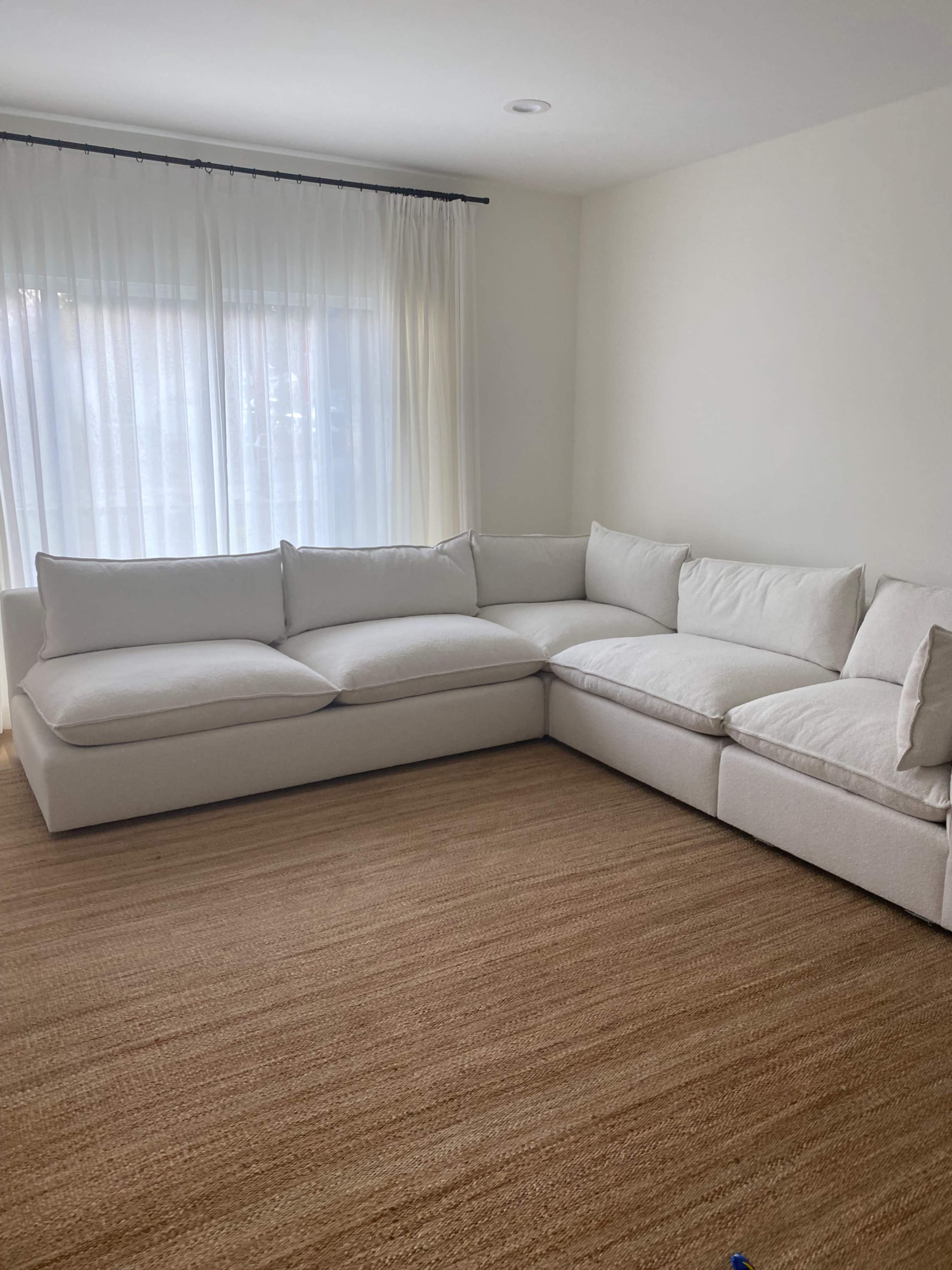 A large white sectional sofa is positioned in a well-lit room with sheer curtains and a natural fiber rug.