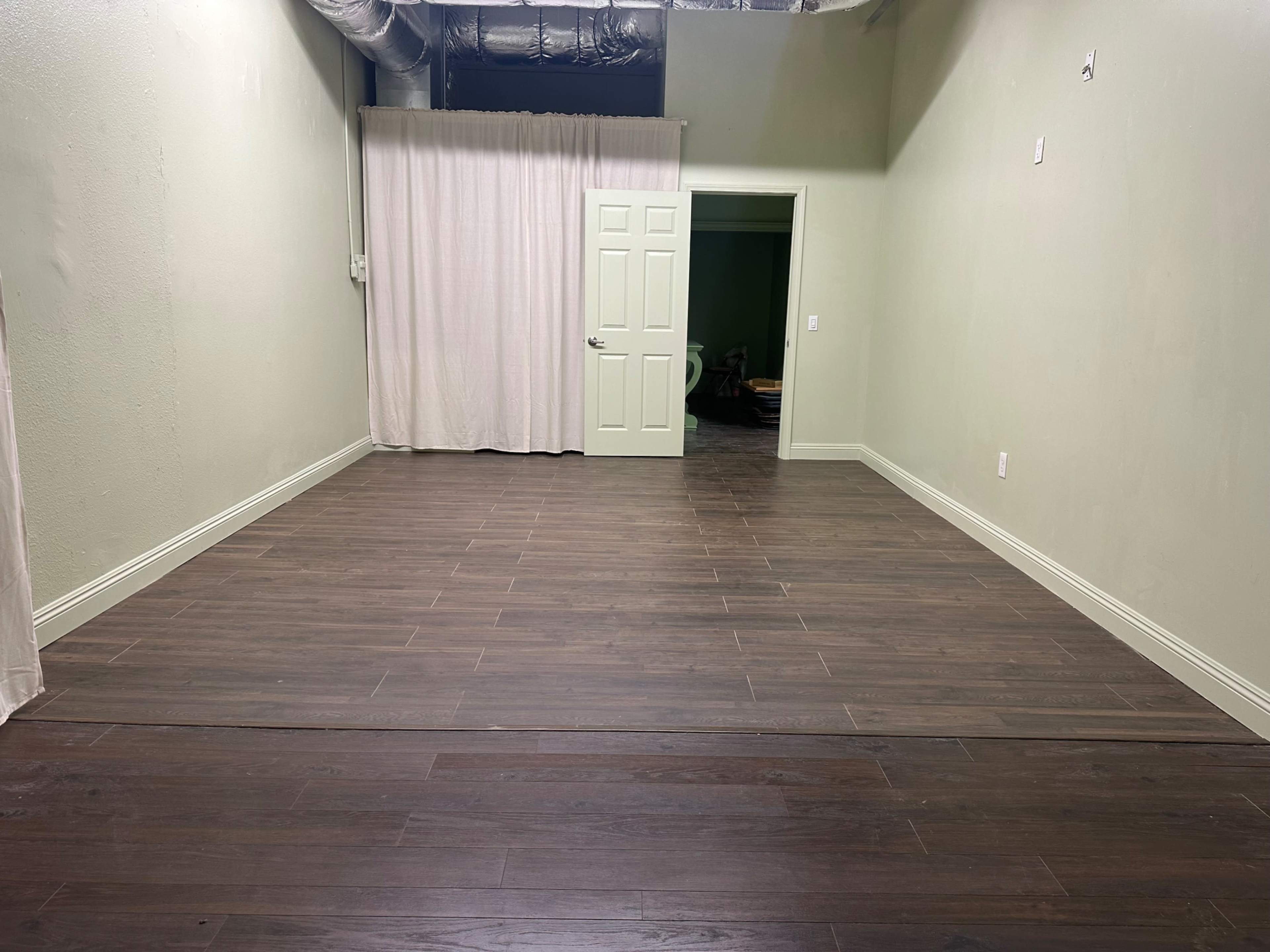The image shows an empty room with wooden flooring, pale green walls, and a white curtain partially covering a door.