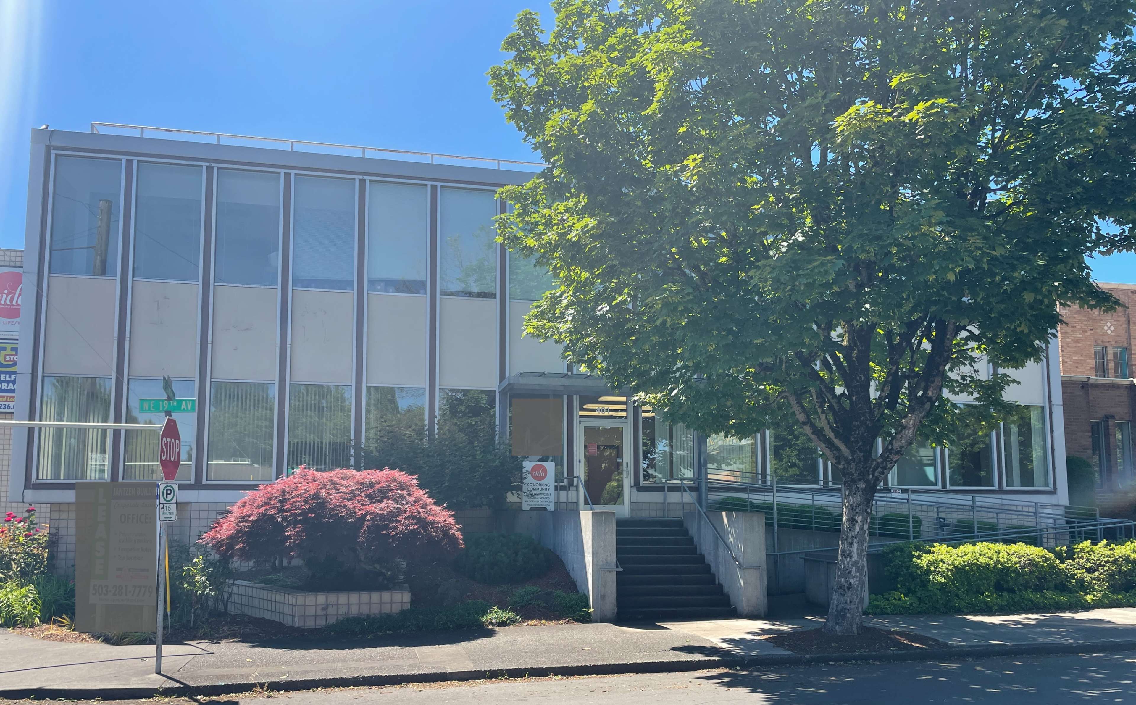 The image shows a two-story building with large windows, a staircase leading to the entrance, and a tree in front, located at a street corner.