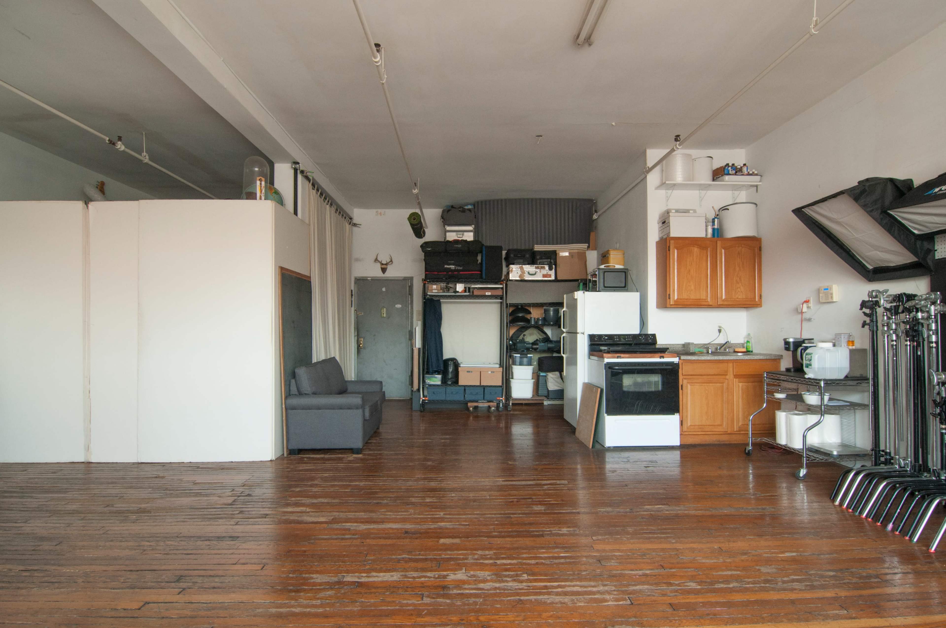 The image shows an open loft space featuring wooden floors, a kitchen area with wooden cabinets and a stove, a gray couch, and various storage boxes along with photography equipment.