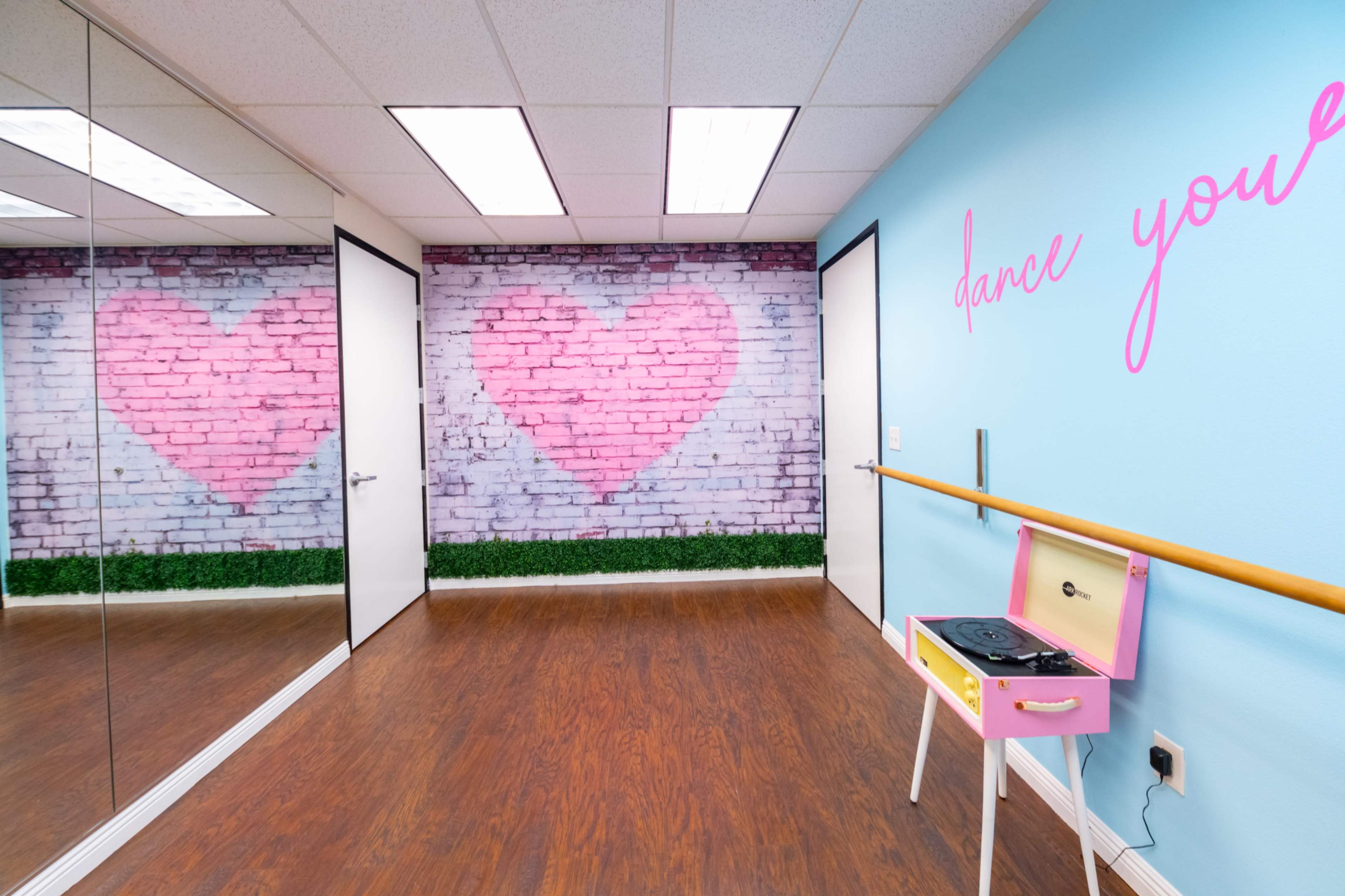 The image shows a dance studio with mirrored walls, a pink heart mural on a brick background, and a pink record player on a ballet bar.