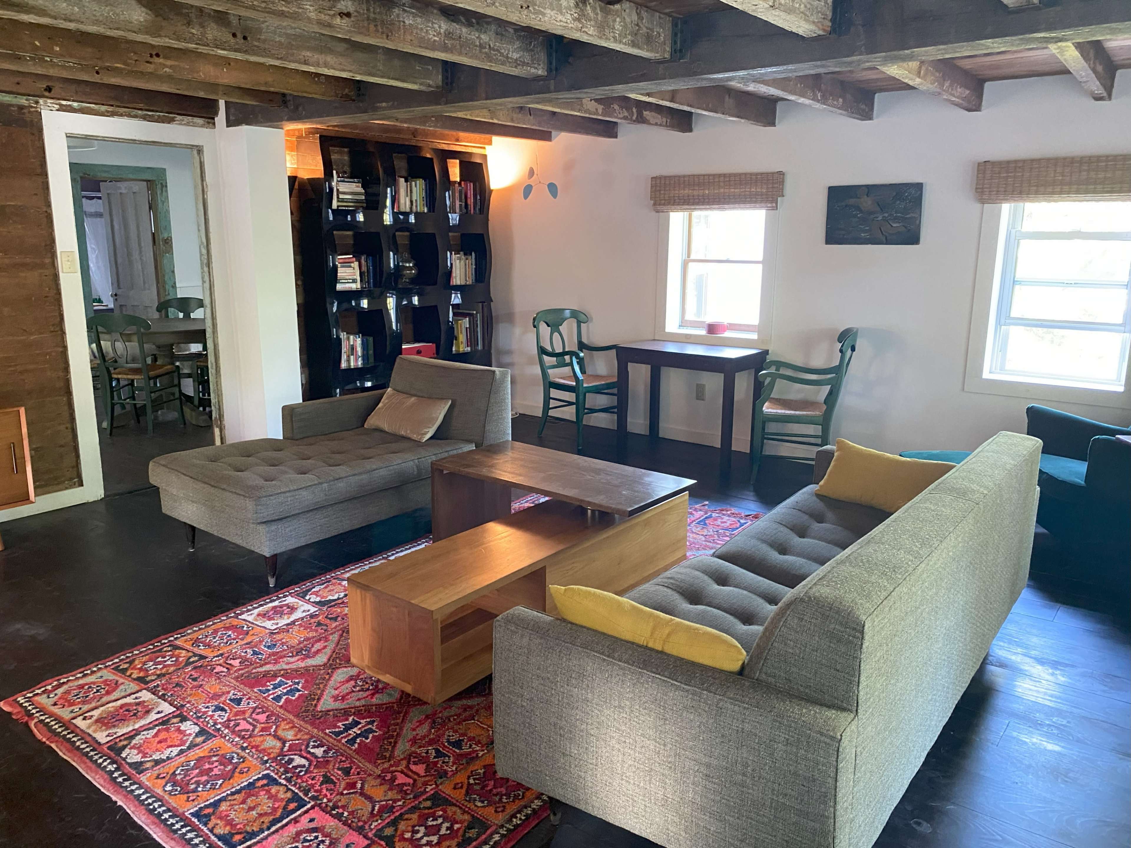 The image shows a cozy living room featuring two couches, a coffee table, and a bookshelf, with natural light streaming through the windows.
