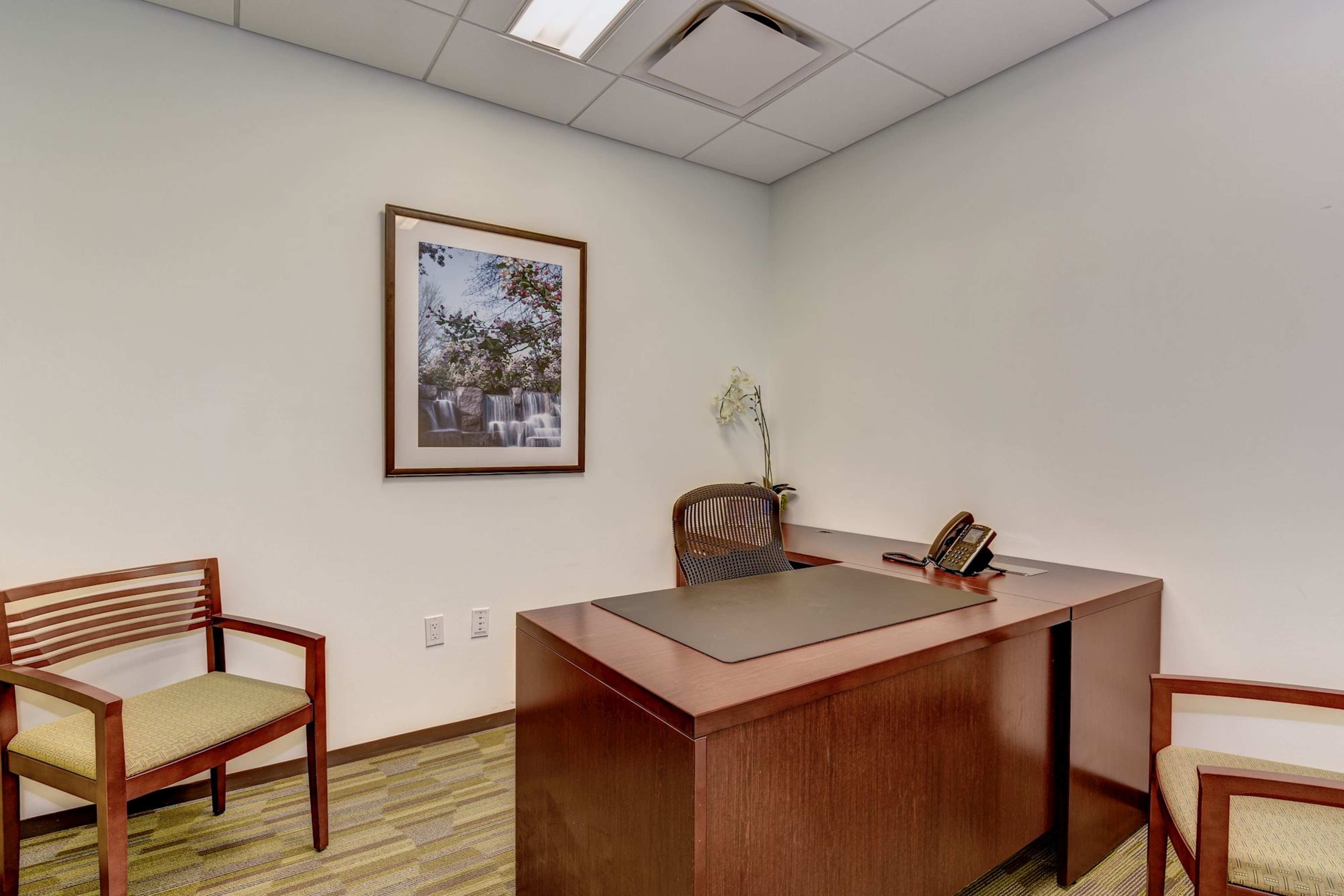 The image shows a simple office space with a wooden desk, an ergonomic chair, two wooden chairs, and a wall-mounted framed picture.