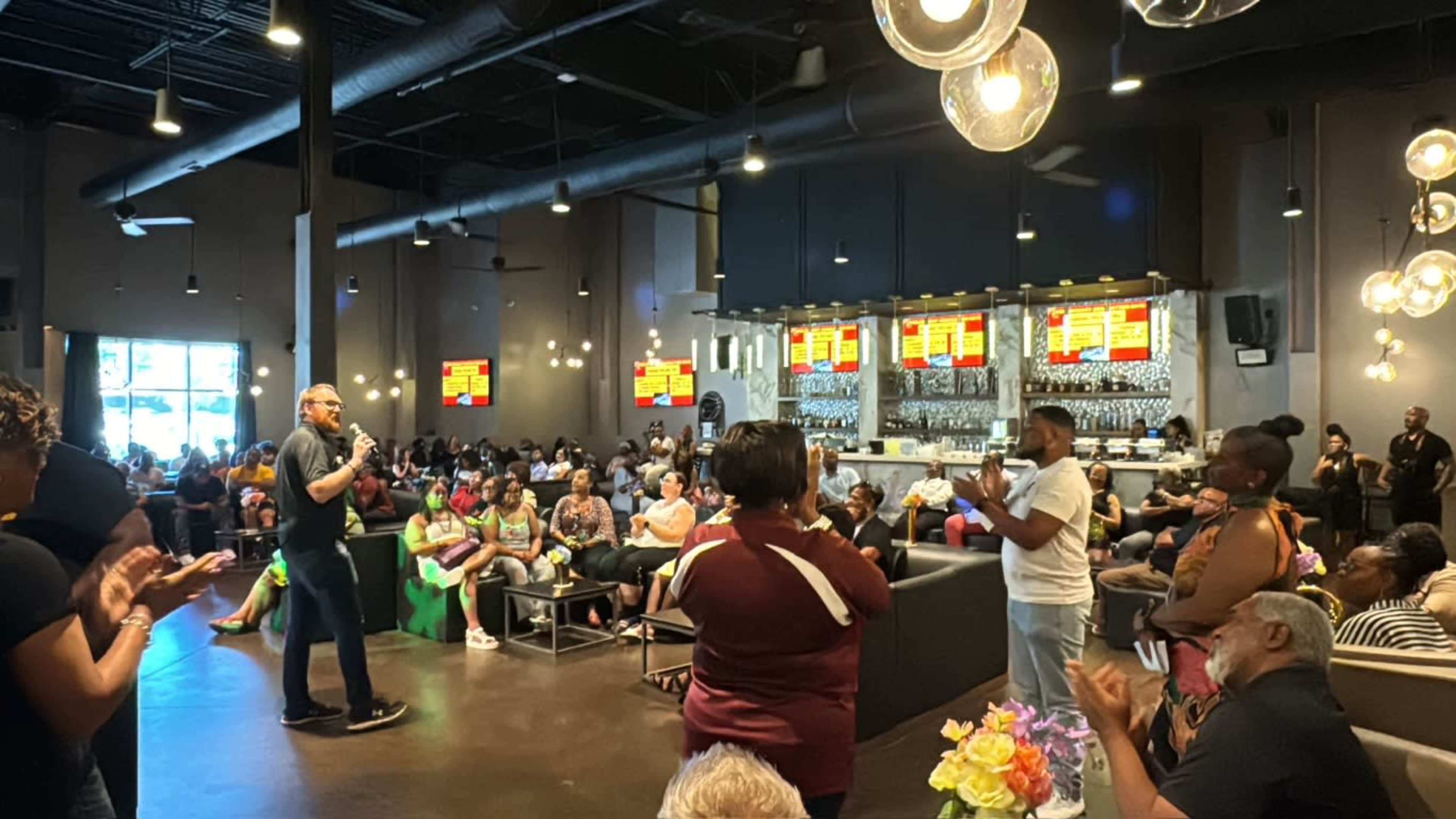 A speaker stands in front of a large audience in a modern event space with a bar in the background.