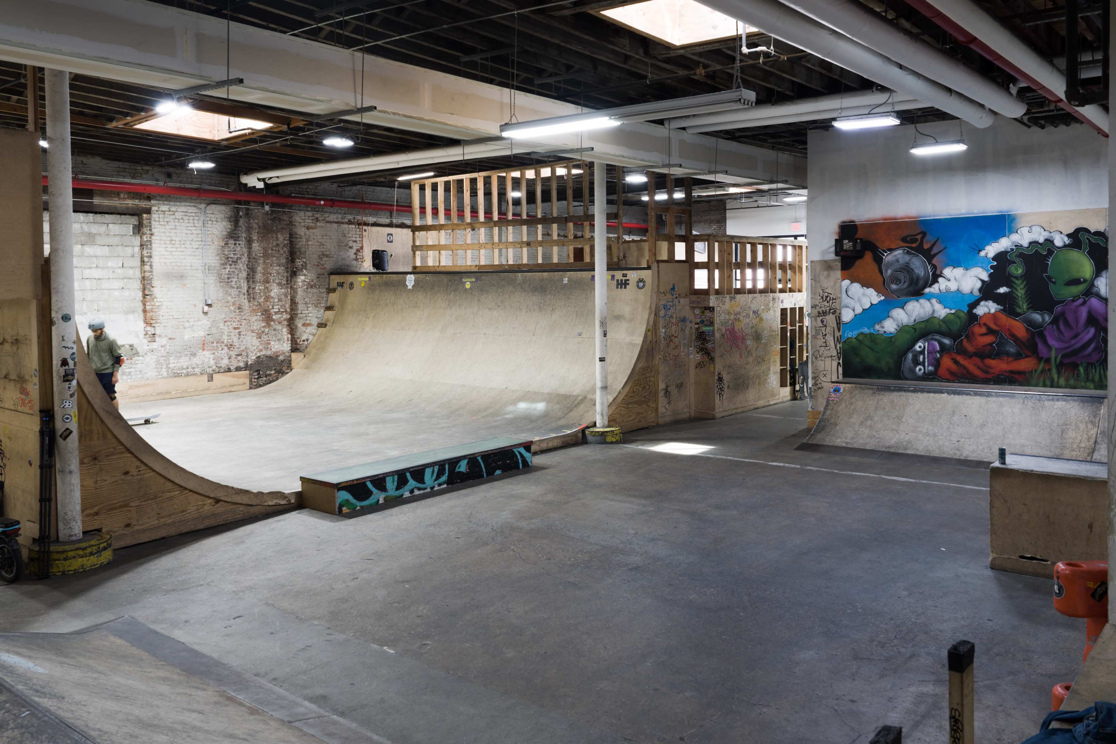 An indoor skatepark with a wood ramp, a half-pipe, and a mural on the wall.